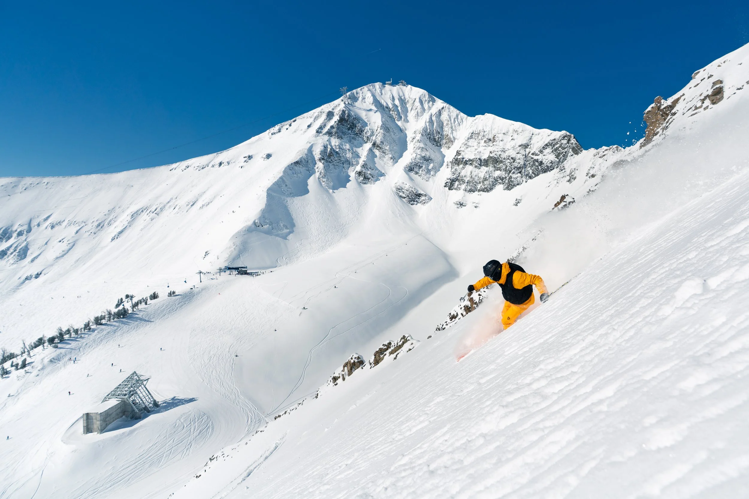 A skier wearing yellow clothing in mid-action on a snowy mountainside with a large snow-covered mountain and clear blue sky in the background.