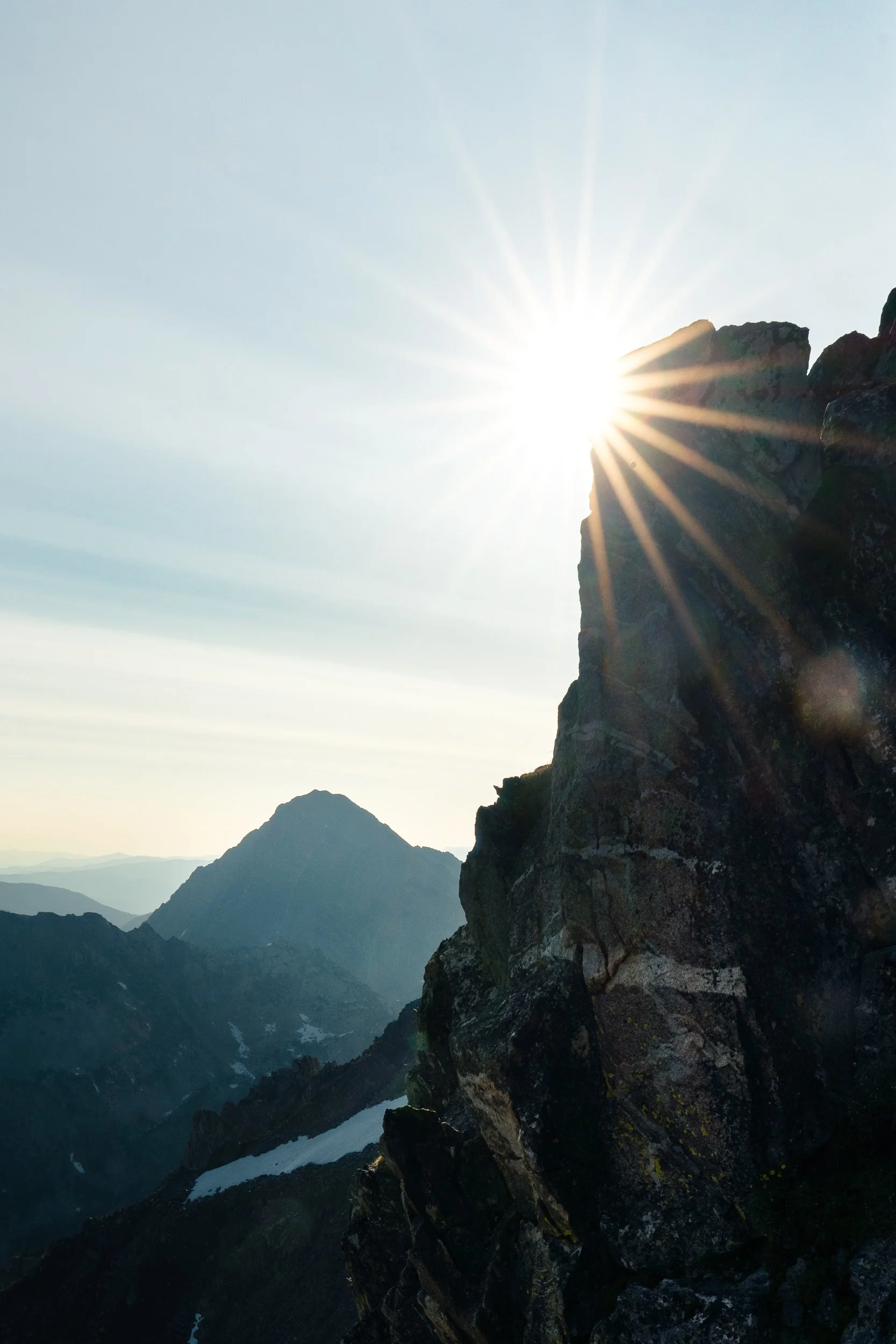 Sun shining behind a mountain, creating sun rays, with multiple mountain peaks and a river or glacier at the bottom.
