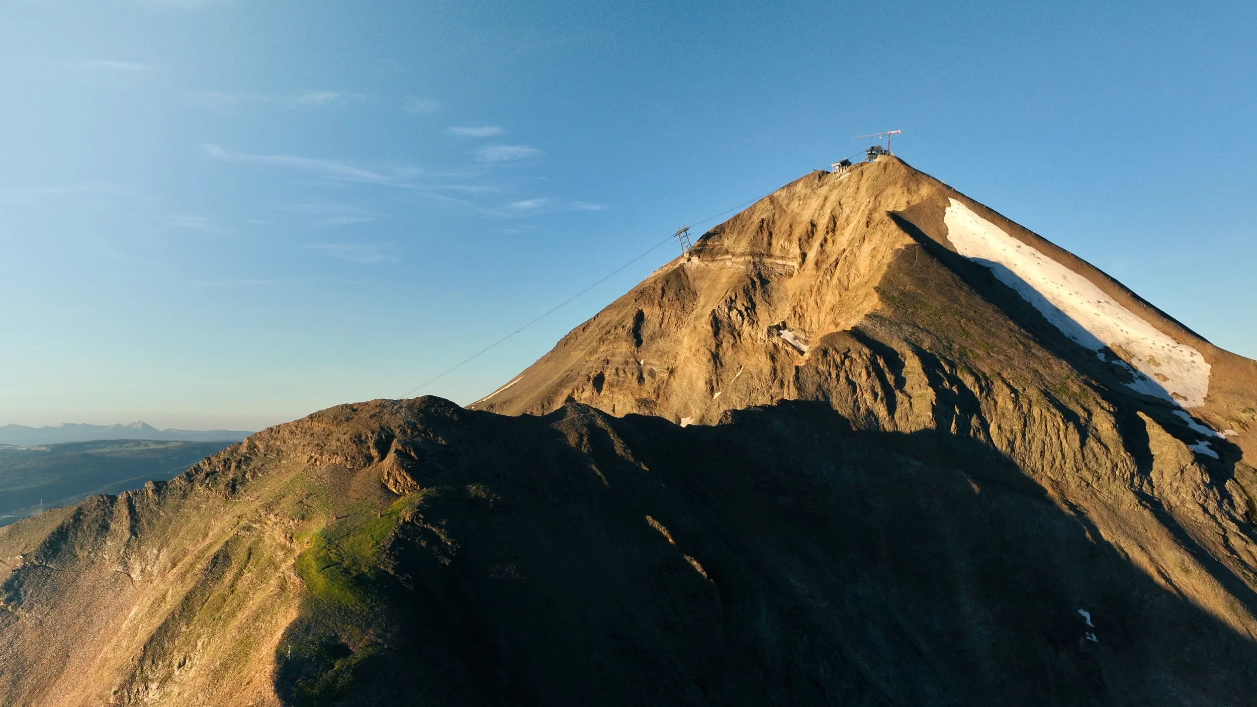 A rugged mountain with steep slopes and patches of snow, under a clear blue sky, with a ski lift at the peak.