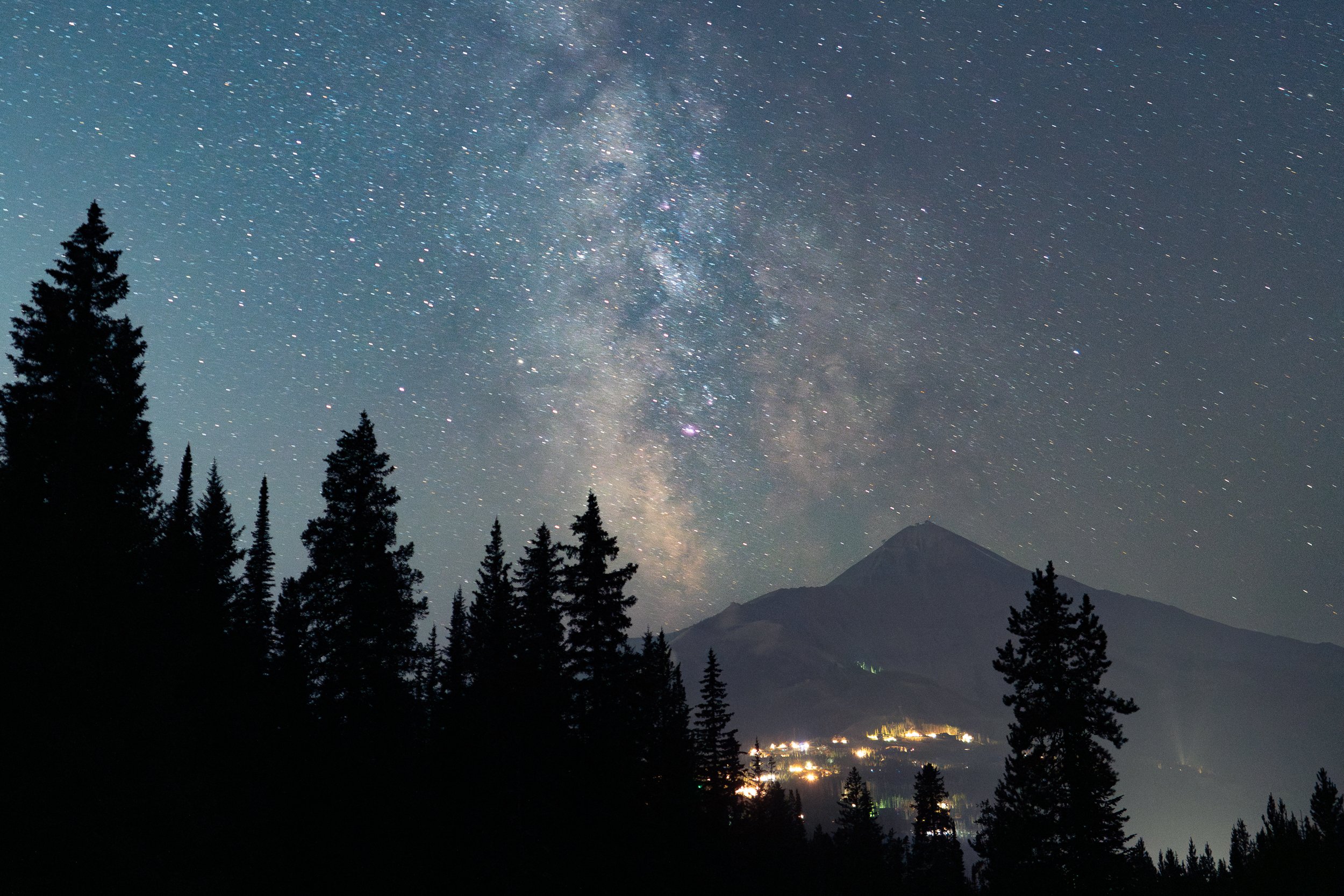 Night sky filled with stars, including the Milky Way galaxy, above a silhouette of tall pine trees and distant mountain with a small village illuminated at the base.