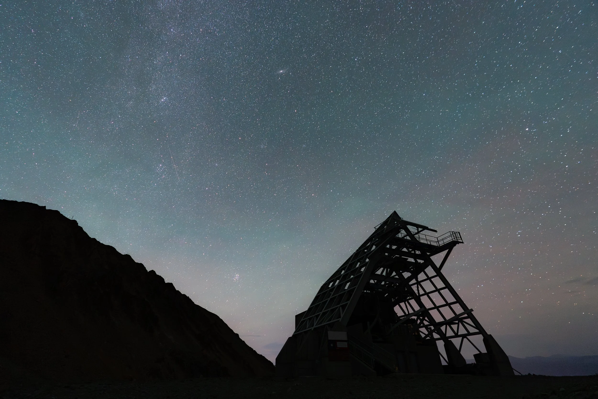 Night sky filled with stars above a dark mountain landscape, with a mountain observatory or telescope structure in the foreground.