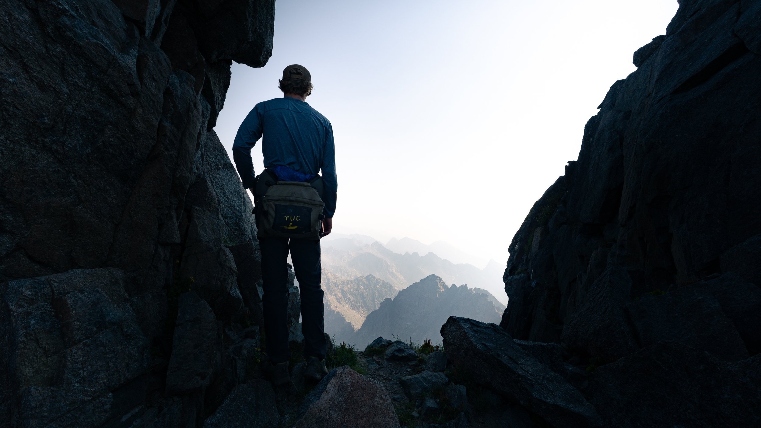 Person standing between rocky mountain cliffs overlooking a mountainous landscape, wearing a cap and a blue jacket with a bag, during daytime.