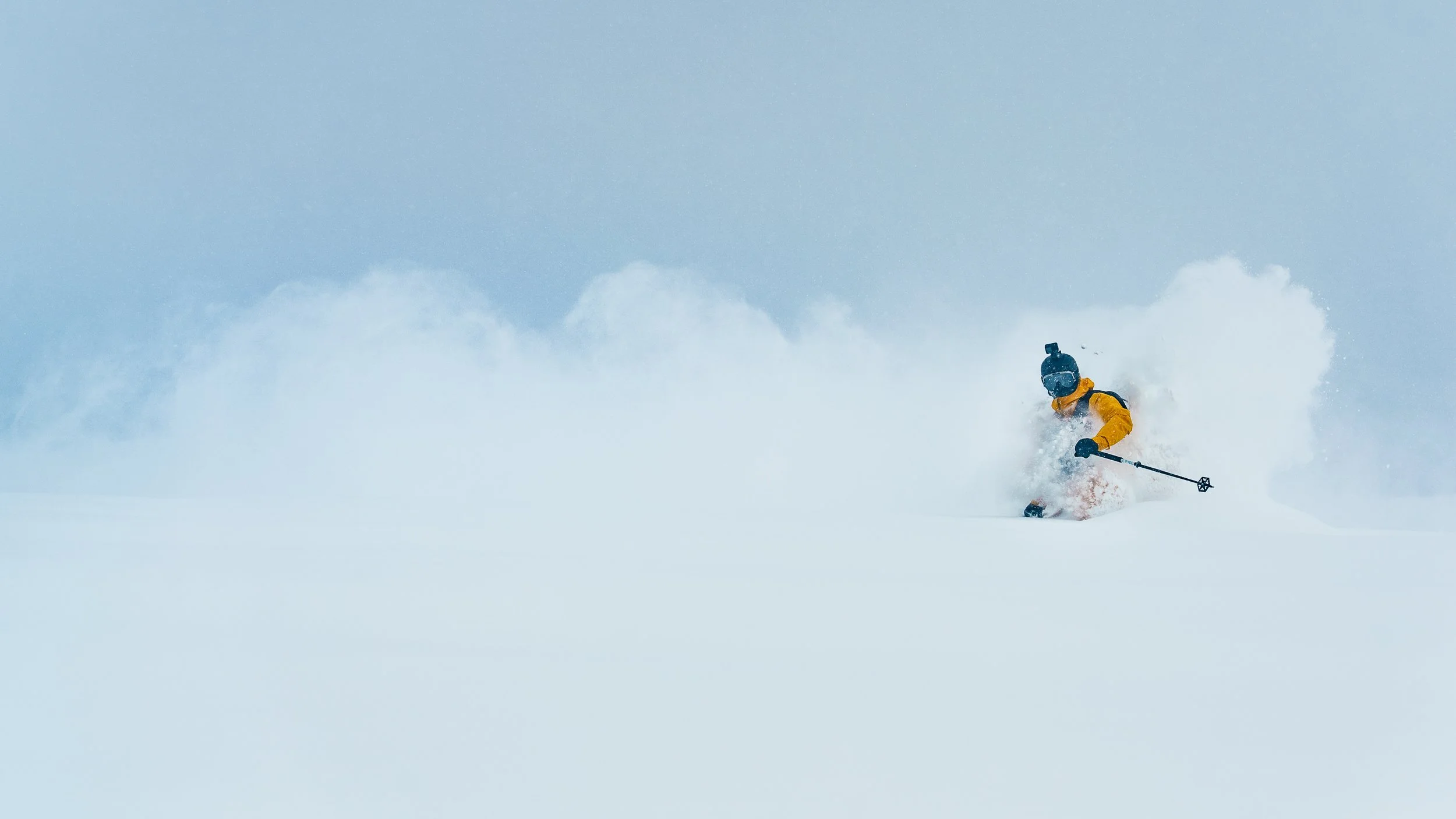 A person wearing a yellow jacket and helmet skiing through deep snow, creating a spray of snow around them against a light blue sky.