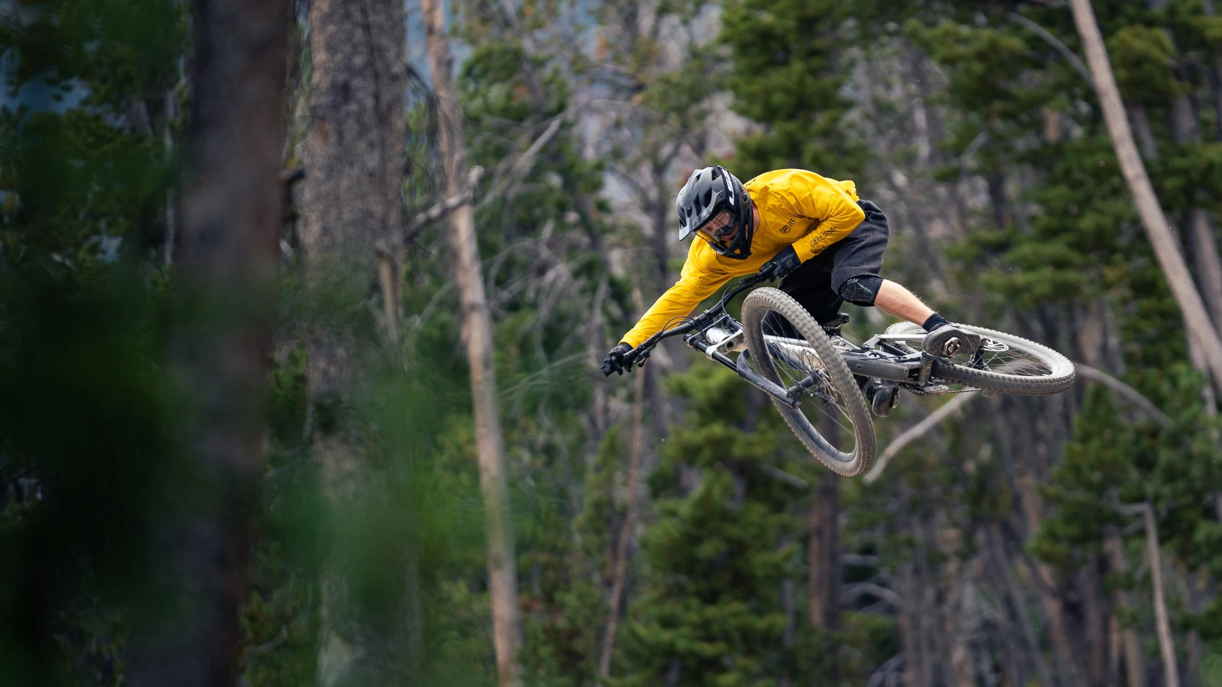 A mountain biker wearing a yellow jersey and black helmet performing a jump or trick in a forested area.