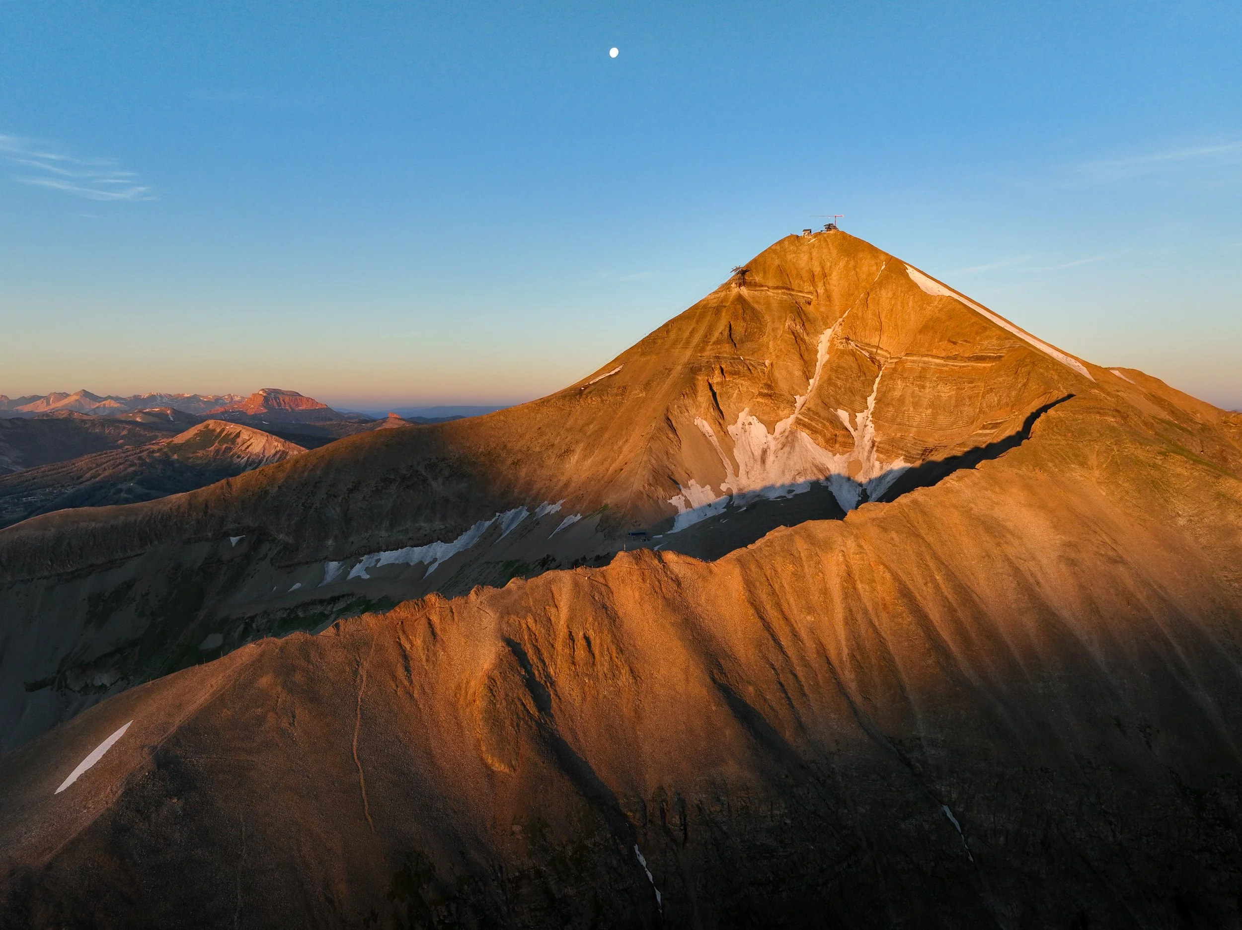 A mountain with patches of snow at sunset or sunrise, with a blue sky and the moon visible.