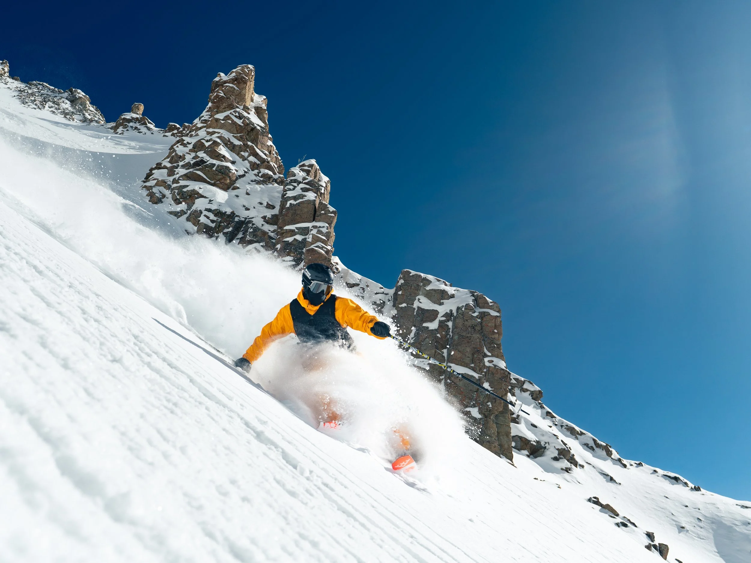 A skier wearing an orange jacket and helmet skiing down a snowy mountain slope with rocky formations and a clear blue sky.