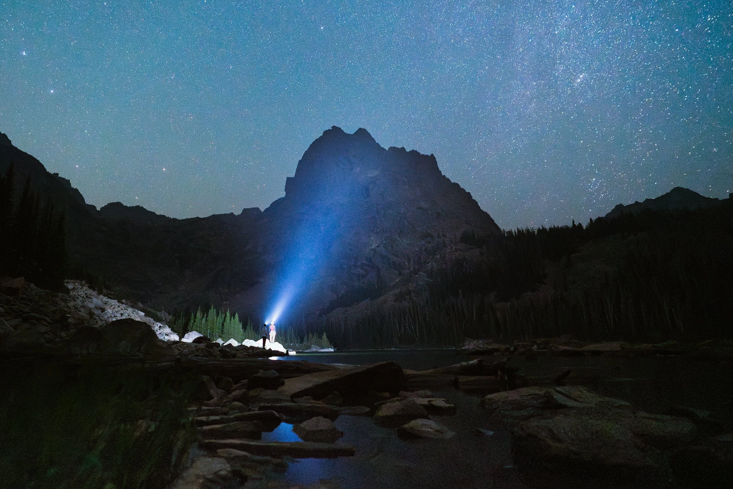 Nighttime landscape of a mountain with a star-filled sky in the background, a blue beam of light shining from a person near the water, and surrounding trees and rocky terrain.