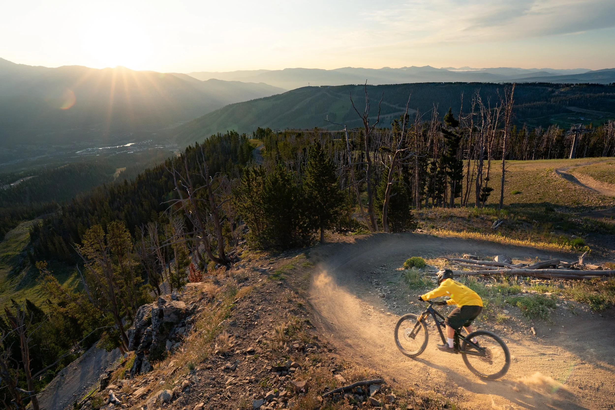A mountain biker wearing a yellow jersey riding on a dirt trail on a mountainside, with a scenic view of layered mountain ranges in the background at sunrise.
