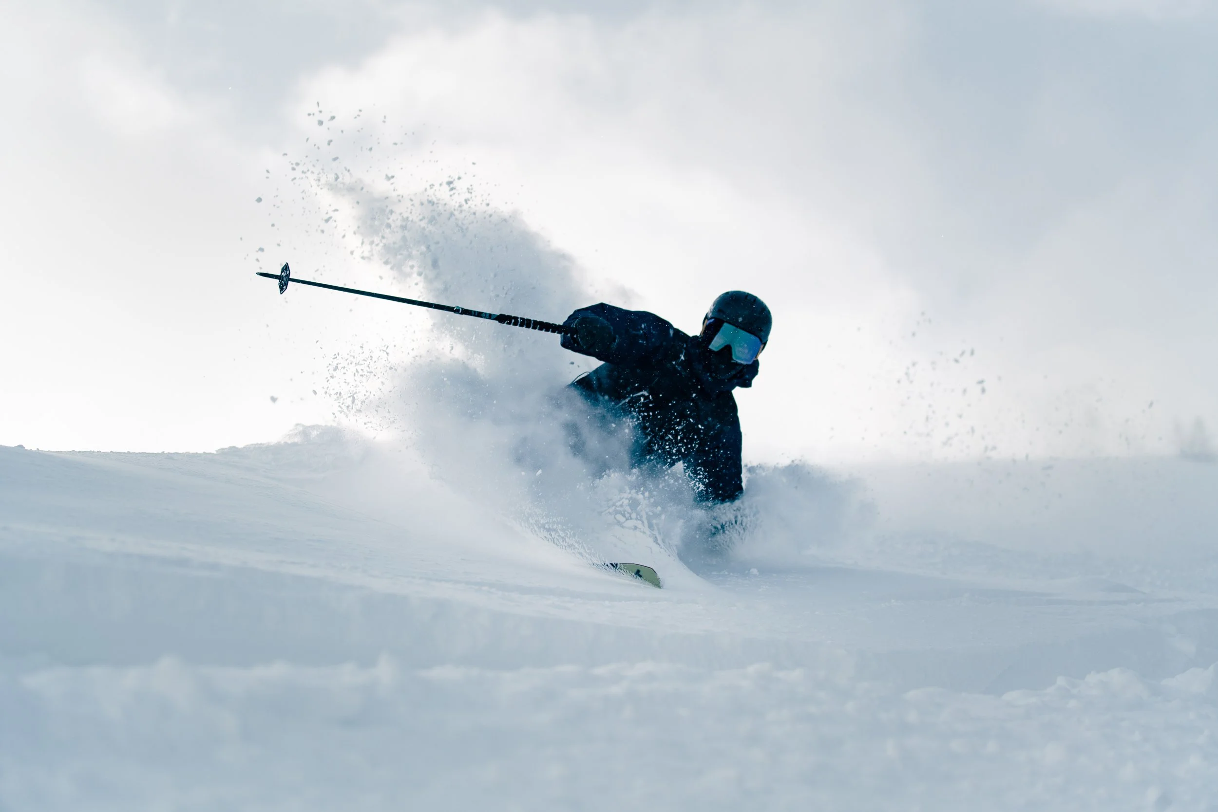 A skier dressed in dark winter gear, including a helmet, making a sharp and deep turn on a snowy bank, kicking up snow and creating a spray of powder in the air against the sky.