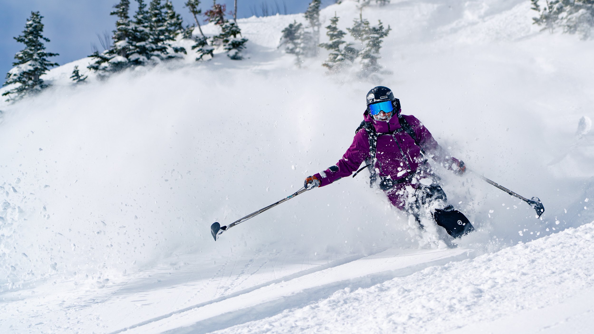 A person wearing a purple jacket, black helmet with blue goggles, and black gloves skiing down a snowy slope surrounded by snow-covered trees.