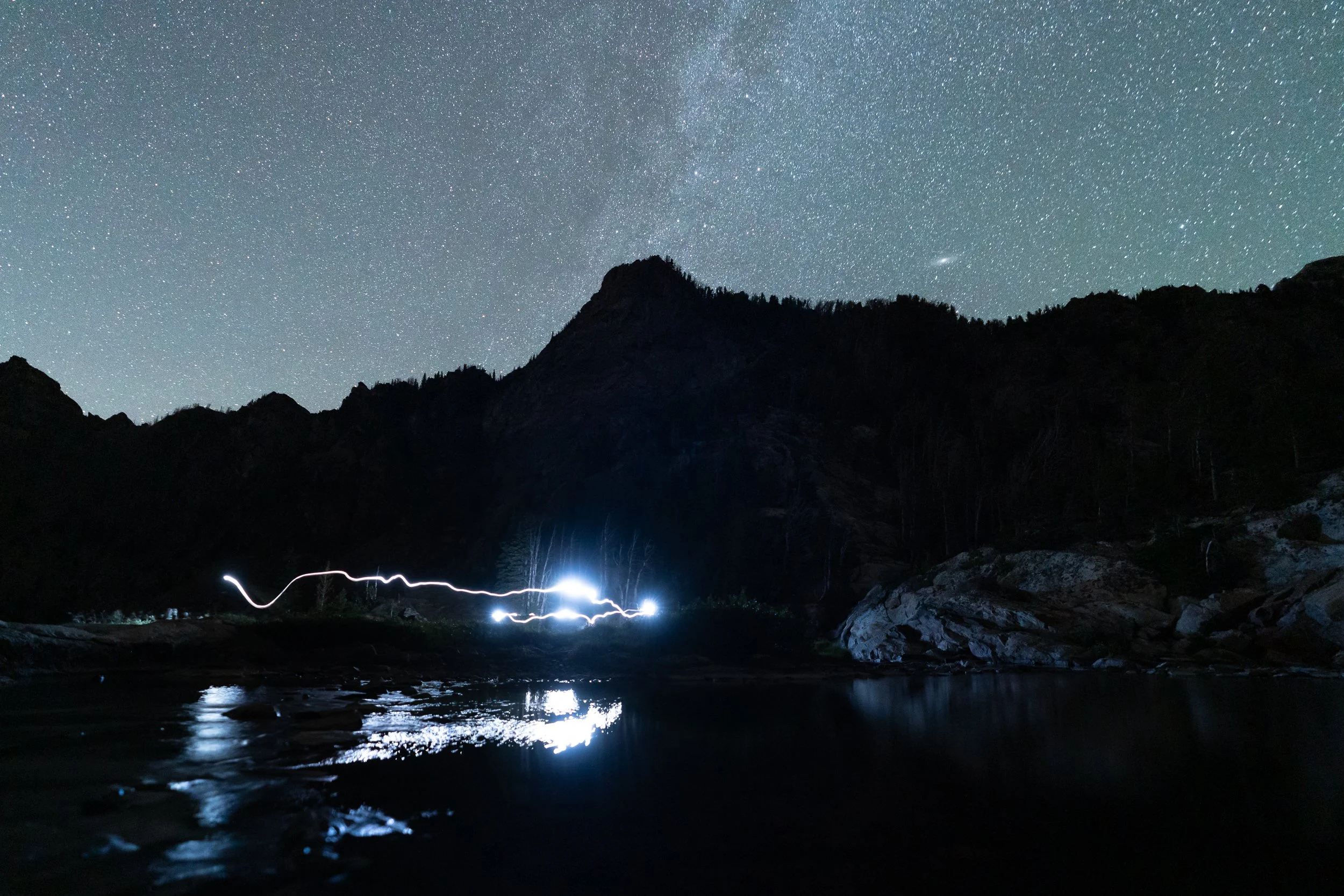 Nighttime landscape with a star-filled sky, mountains silhouette, and a body of water reflecting the stars. There is a light trail near the water's edge.