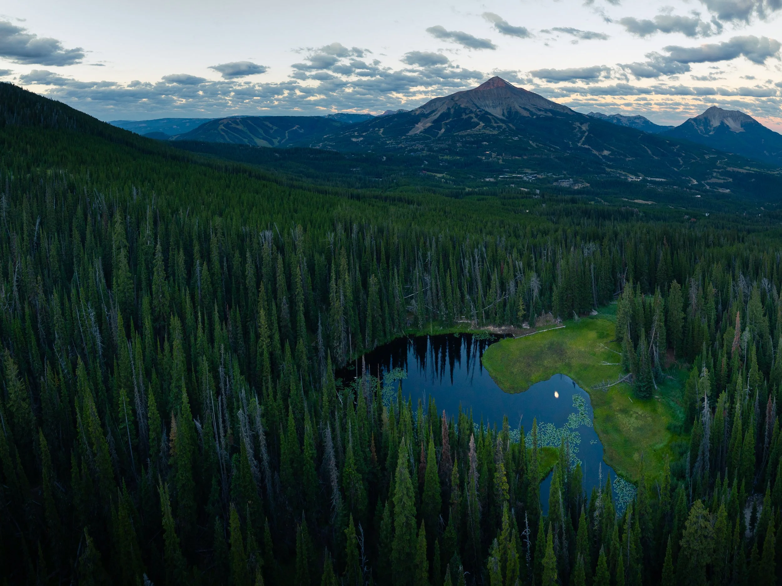A scenic landscape of a mountain with a valley below, surrounded by dense forest and a small body of water, under a partly cloudy sky.