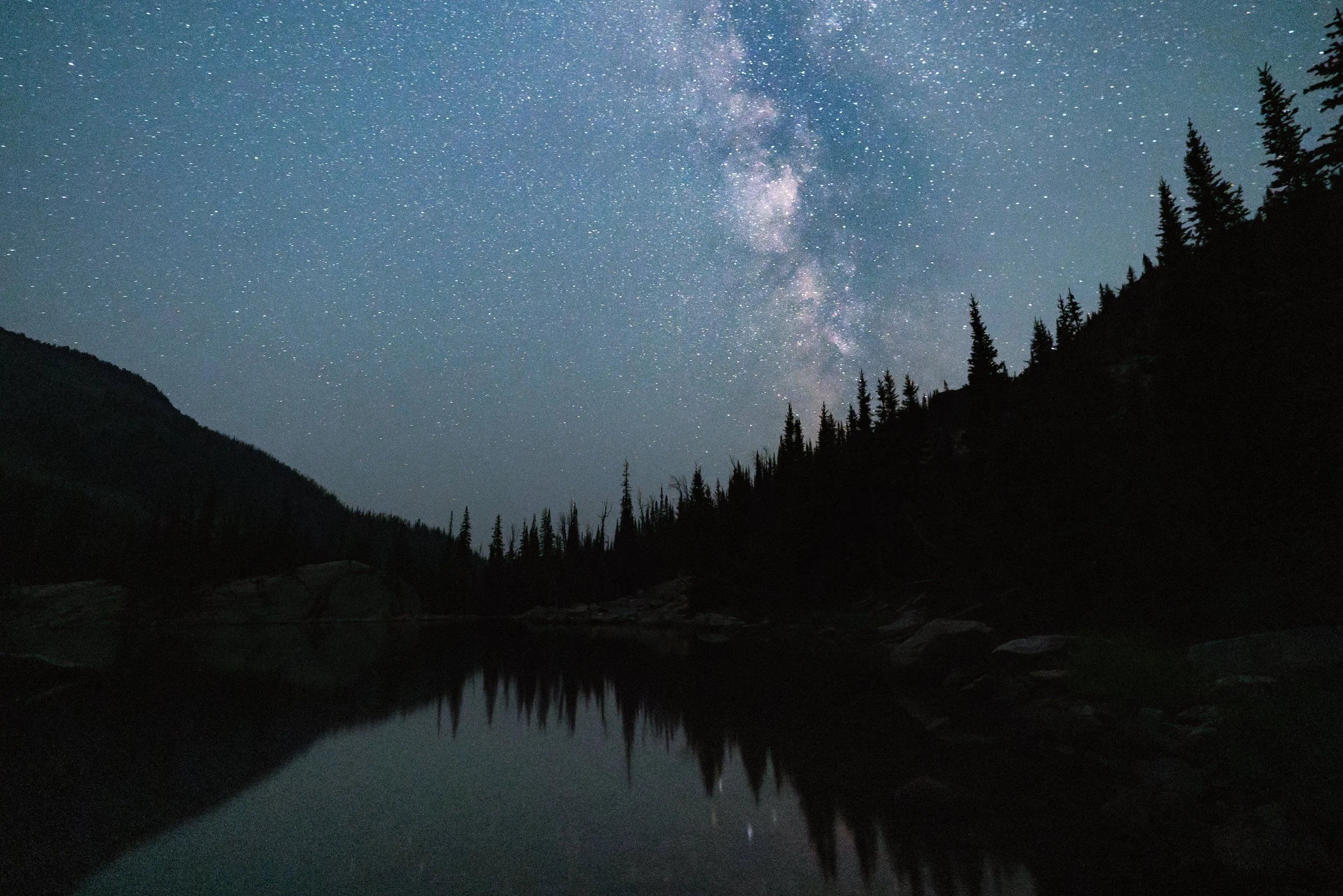 Night sky filled with stars and the Milky Way galaxy, with dark silhouettes of trees and mountainous terrain reflected in a calm lake.