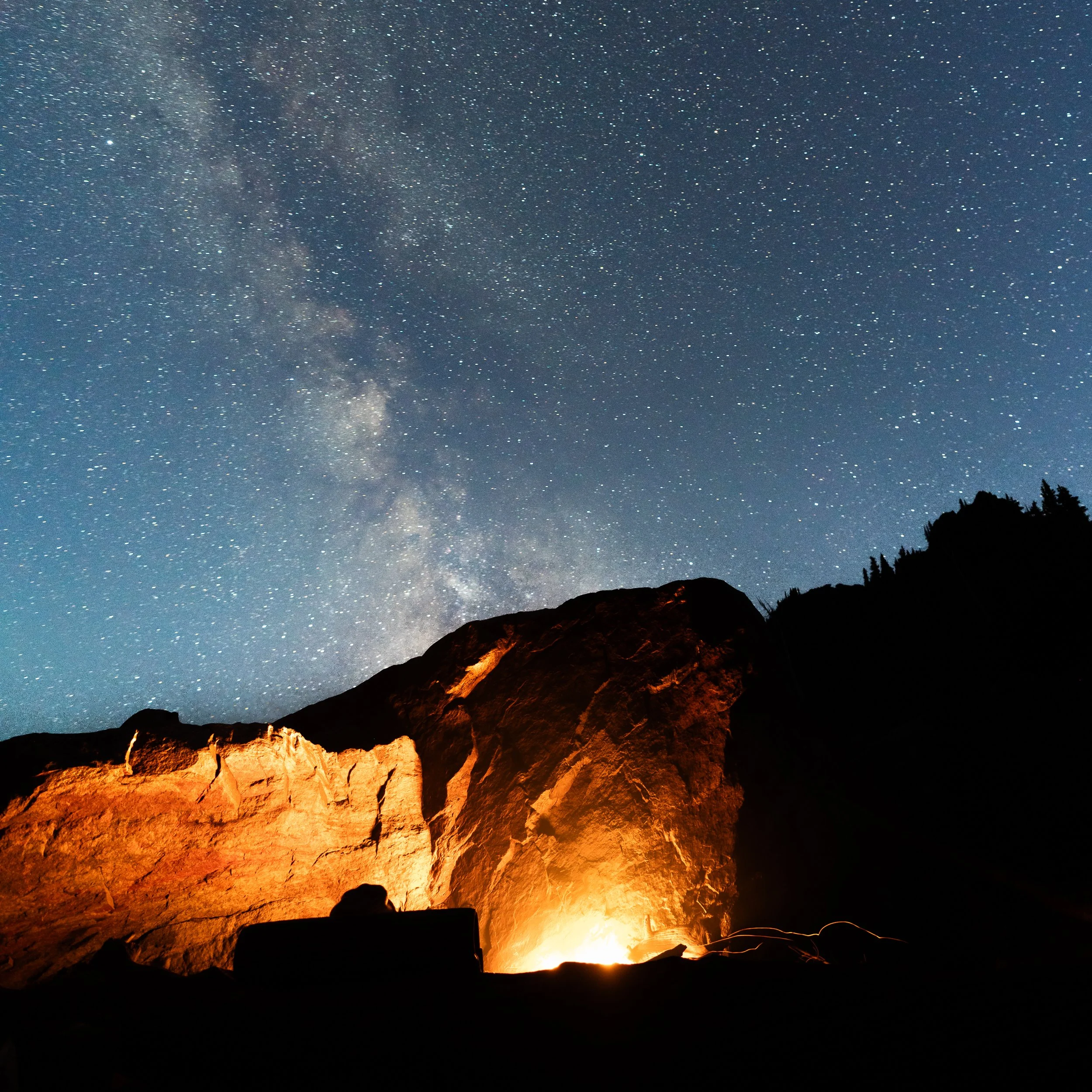 Night sky filled with stars and a faint glimpse of the Milky Way galaxy above a mountain landscape with illuminated rocky cliffs and dark silhouettes of trees in the foreground.