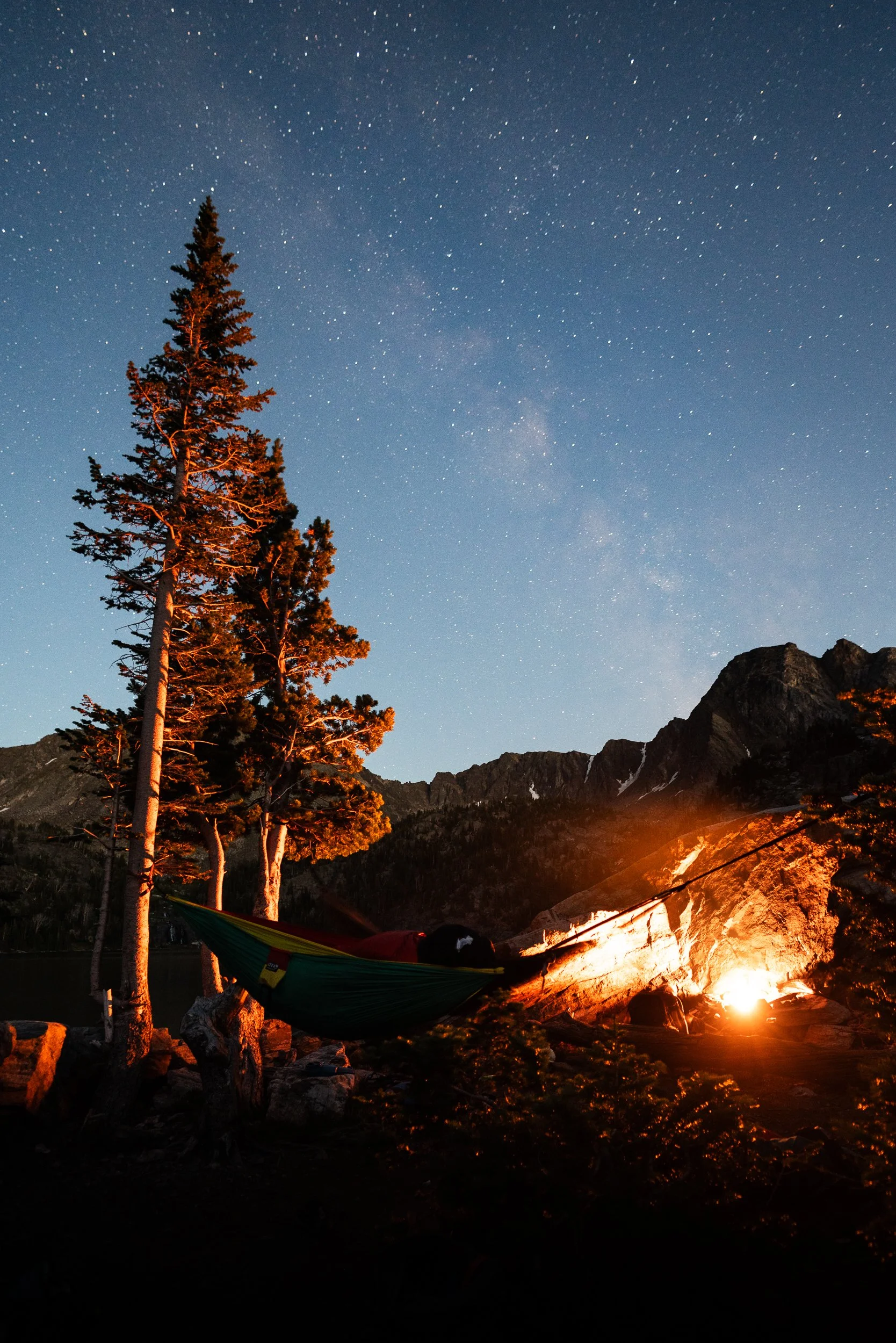Nighttime scene in a mountainous area with a campfire illuminating rocks, trees, and a hammock tied between two trees, under a starry sky.