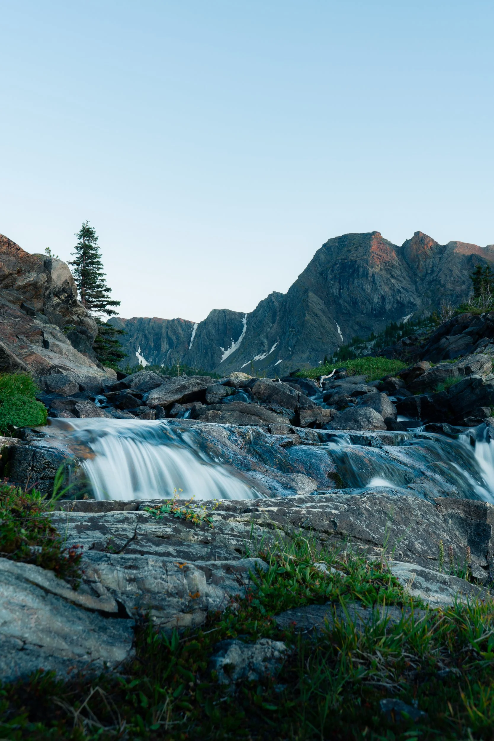 A mountain landscape with rocky terrain, a small waterfall in the foreground, green vegetation, and snow patches on the mountain slopes. Clear sky overhead.