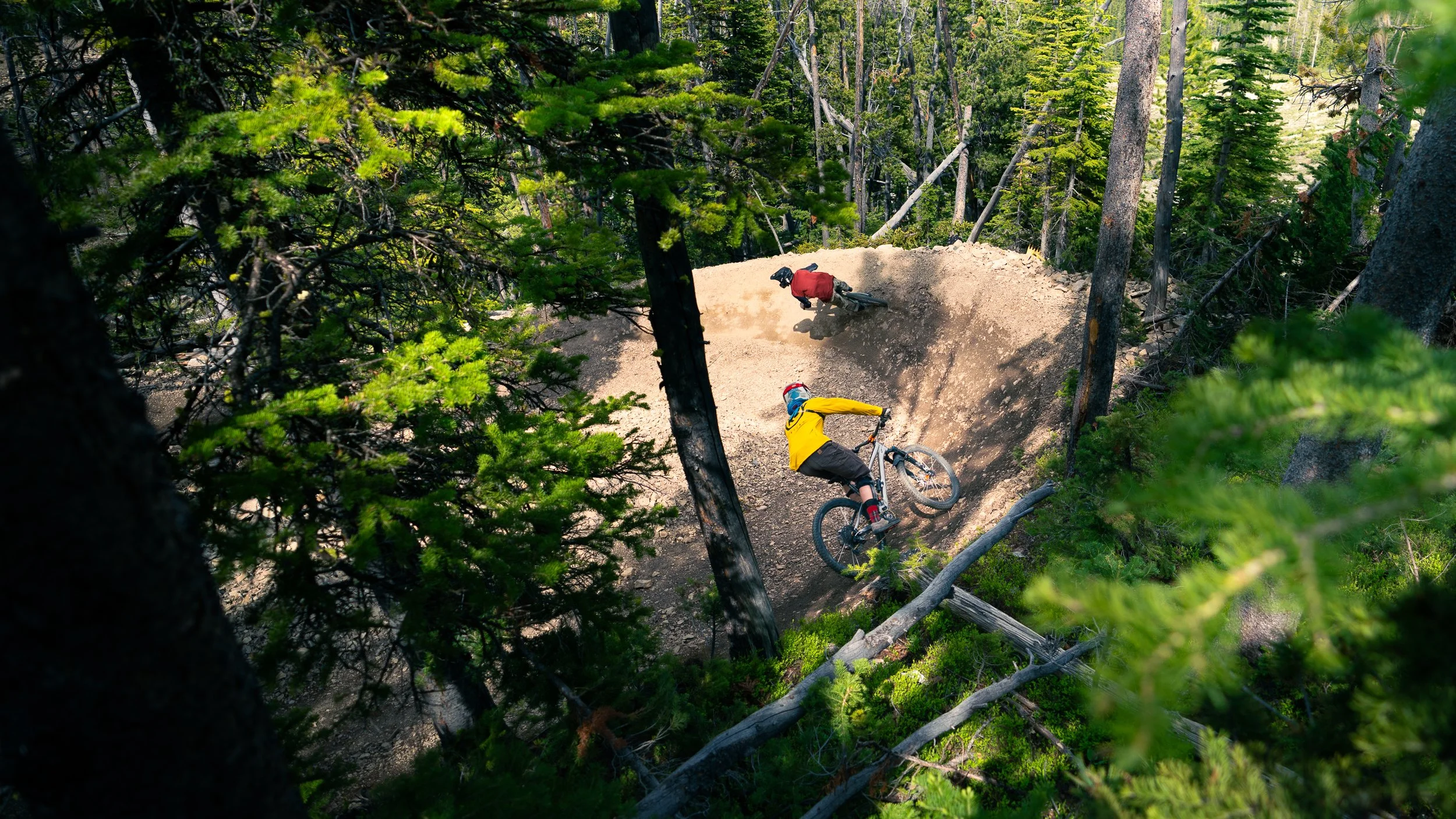 Two mountain bikers riding on a dirt trail through a dense green forest, viewed from above.