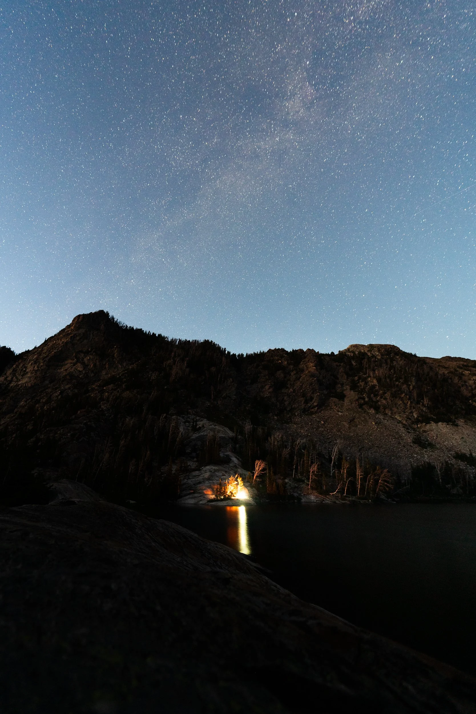 Nighttime landscape with a star-filled sky, mountains, and a lake reflecting a small campfire or light source near the shoreline.