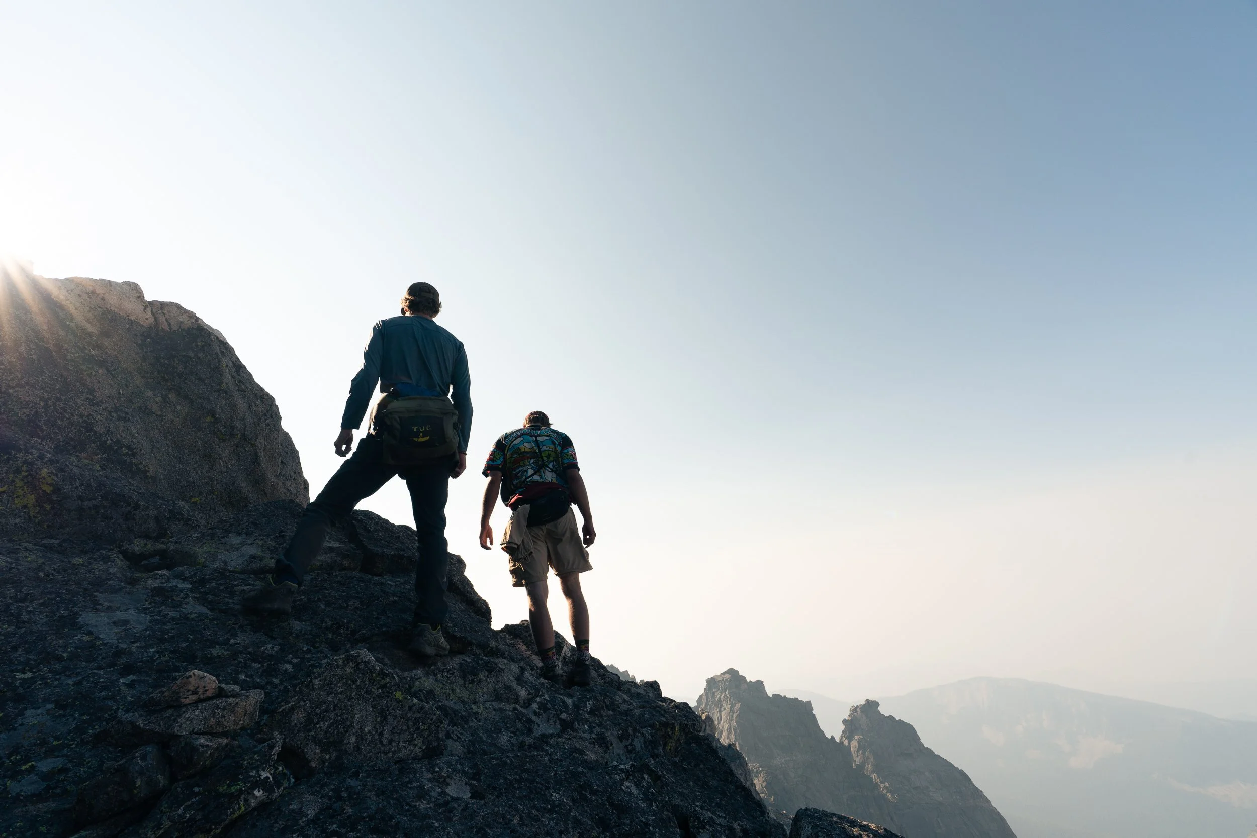Two hikers climbing a rocky mountain trail with a mountain range in the background under a clear sky.