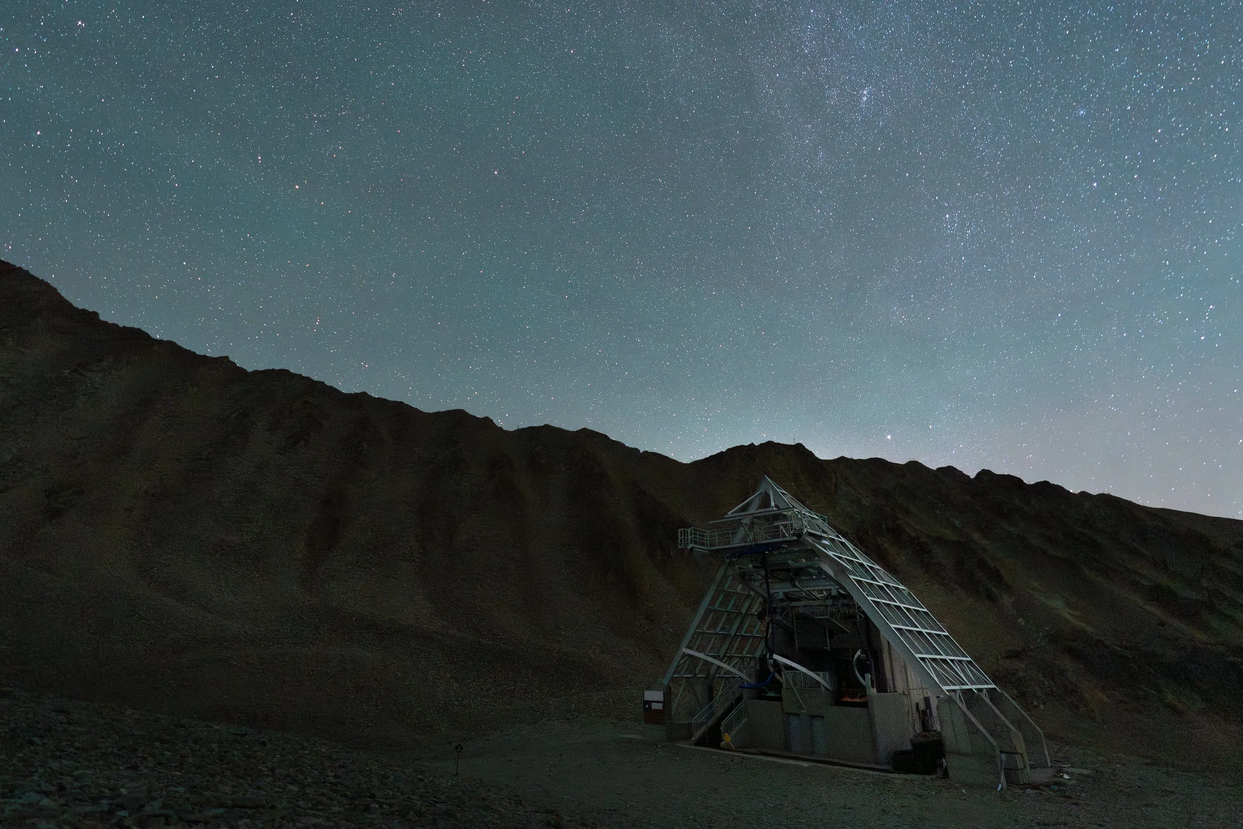 A starry night sky over a mountain landscape with a telescope structure in the foreground.
