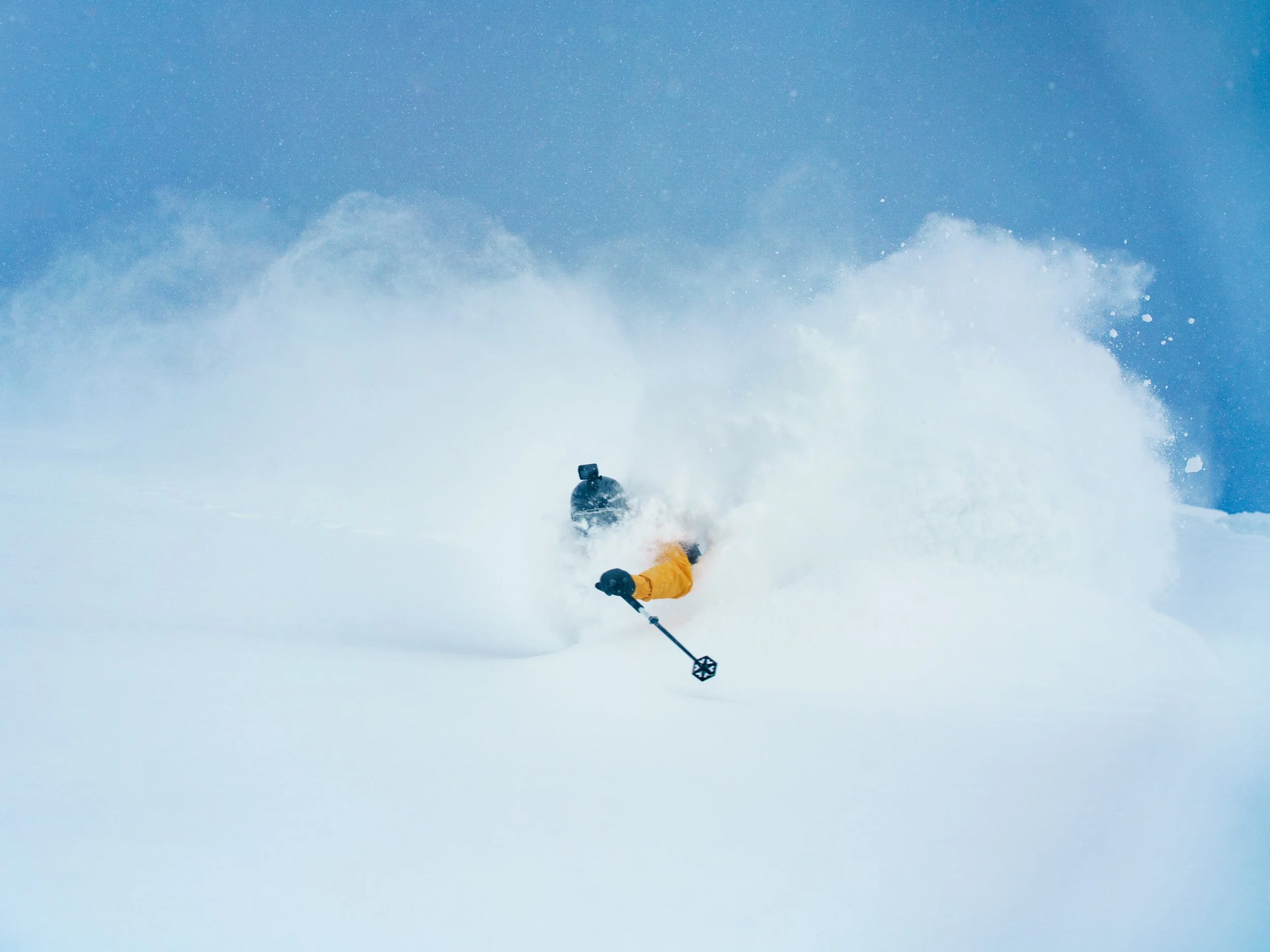 Person skiing downhill in snow, snow spray in the air under clear blue sky.