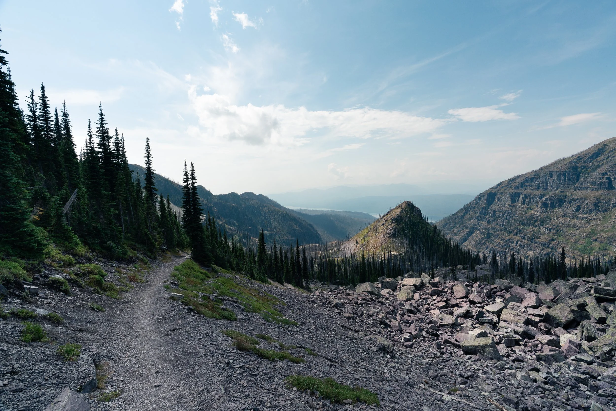 A mountain trail surrounded by pine trees, rocky terrain, and distant mountain ridges under a partly cloudy sky.
