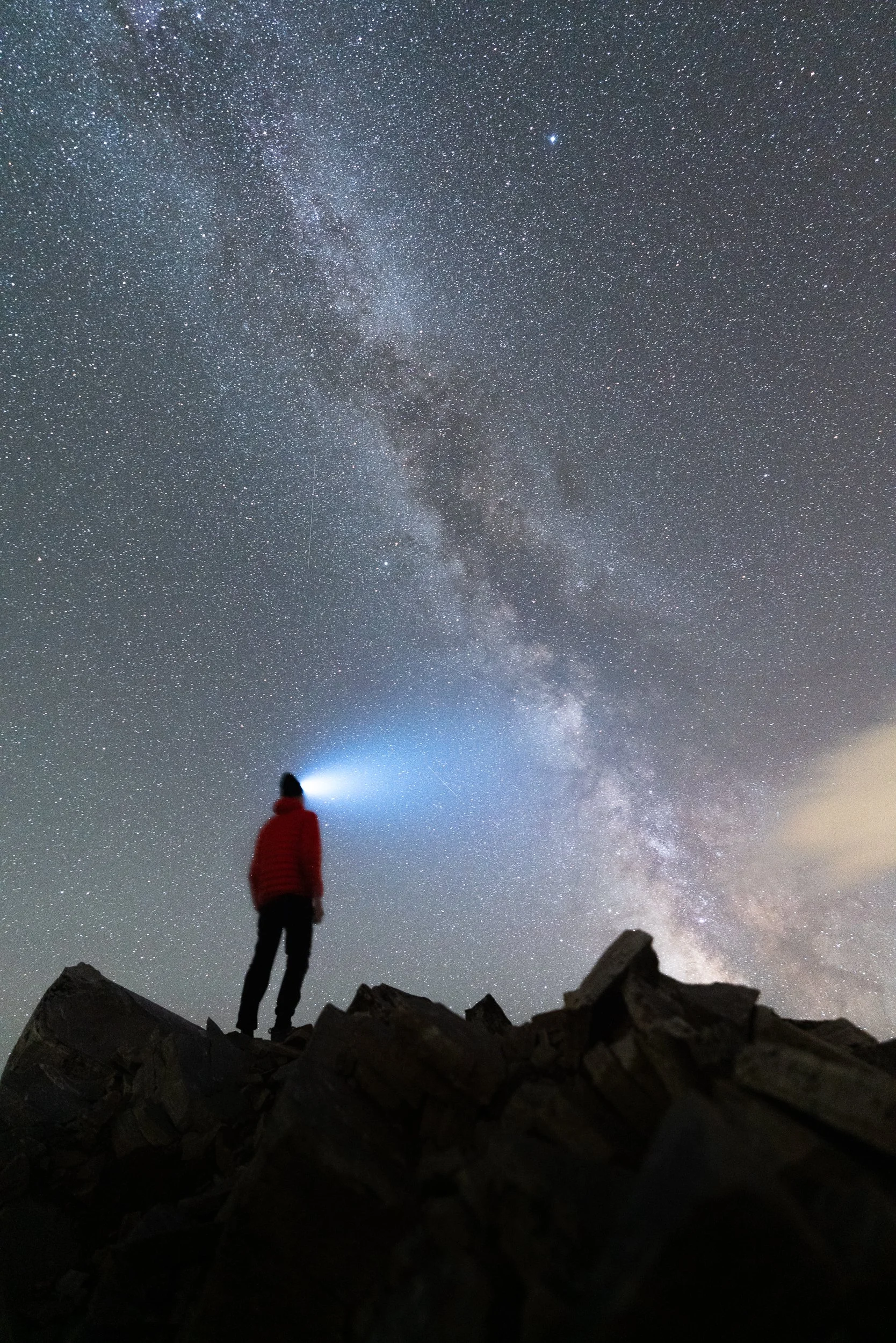 Person standing on rocky terrain at night, looking up at a star-filled sky and the Milky Way galaxy with a flashlight in hand.