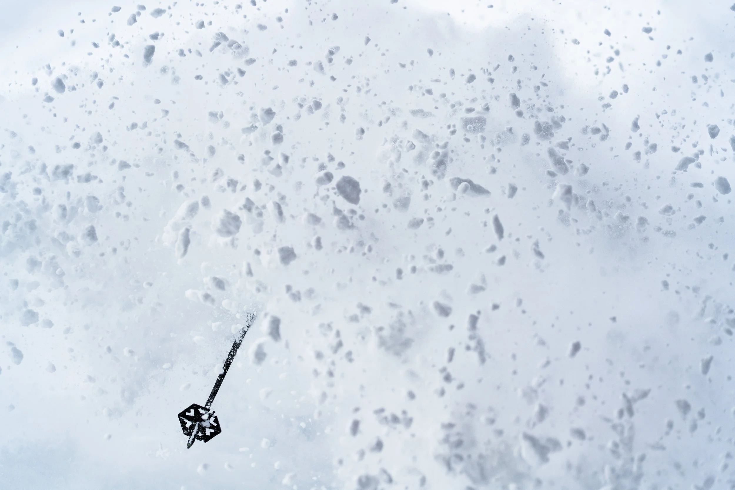 A person holding a ski pole up in snow with snow-covered ground and snowflakes falling, possibly skiing or snowboarding.