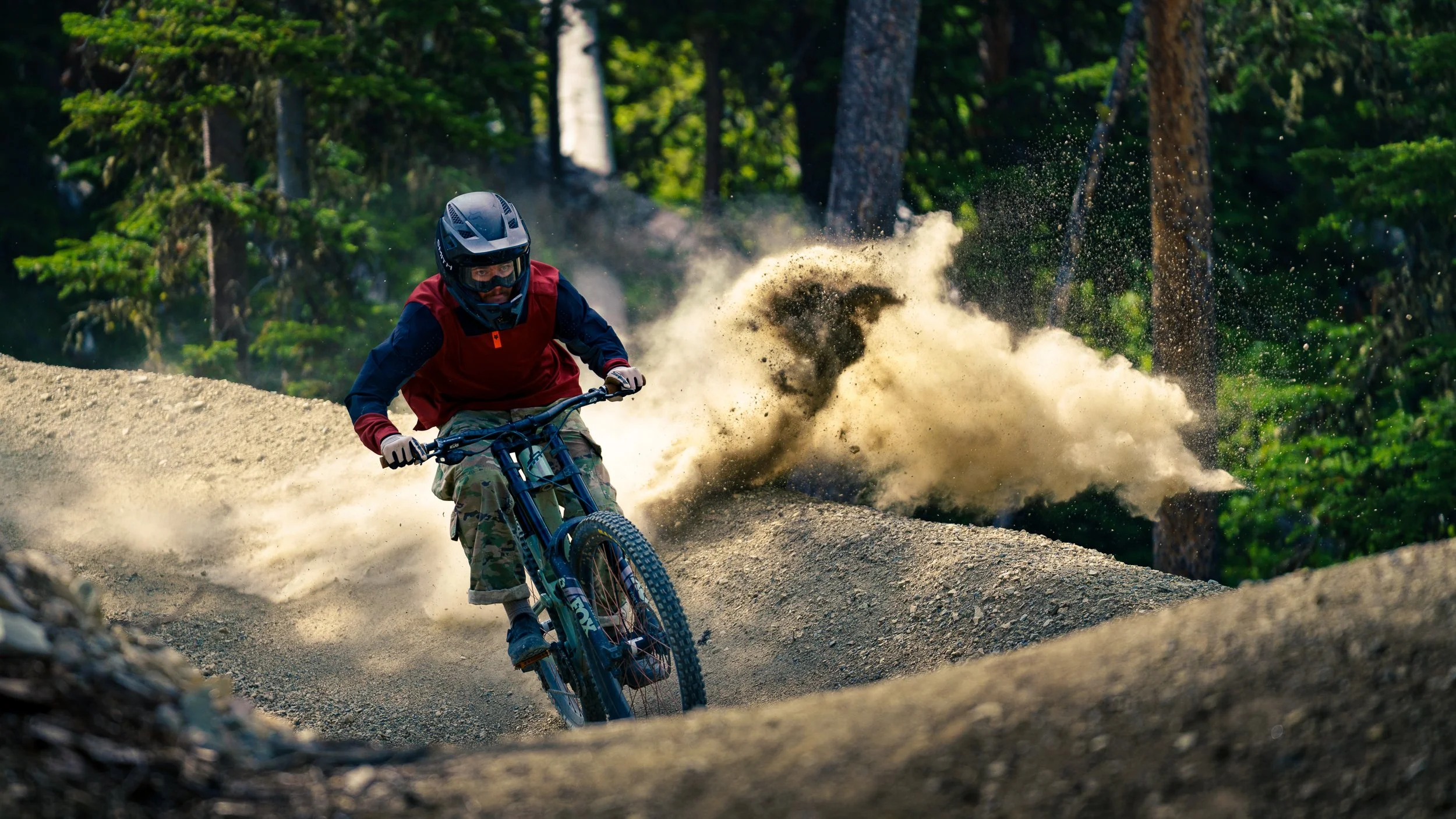 A person riding a mountain bike on a dirt trail in a forest, wearing a helmet, red and black jersey, camouflage pants, and kicking up dust behind them.