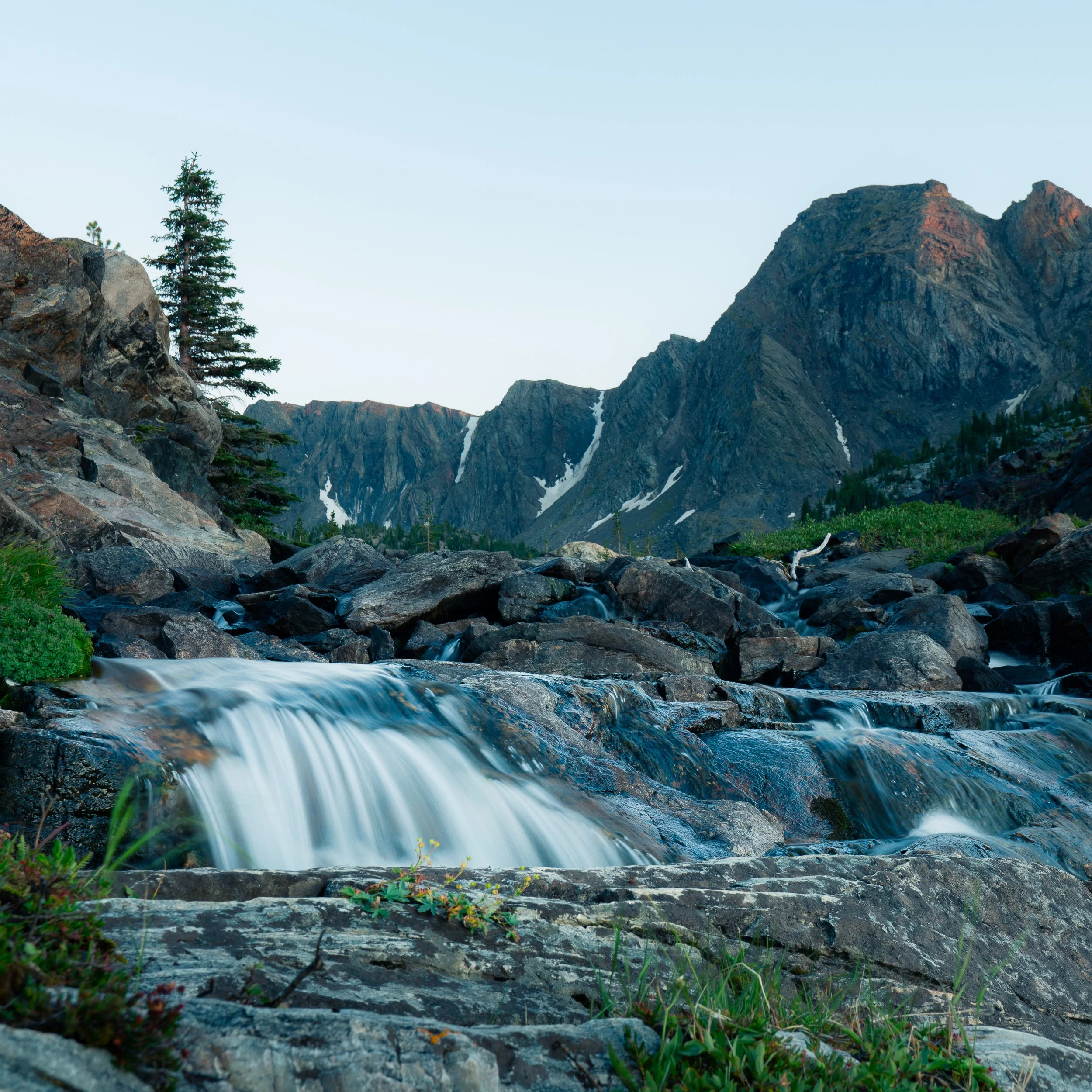 A mountain landscape with a small waterfall flowing over rocks, green vegetation in the foreground, mountain peaks in the background, and a clear sky.