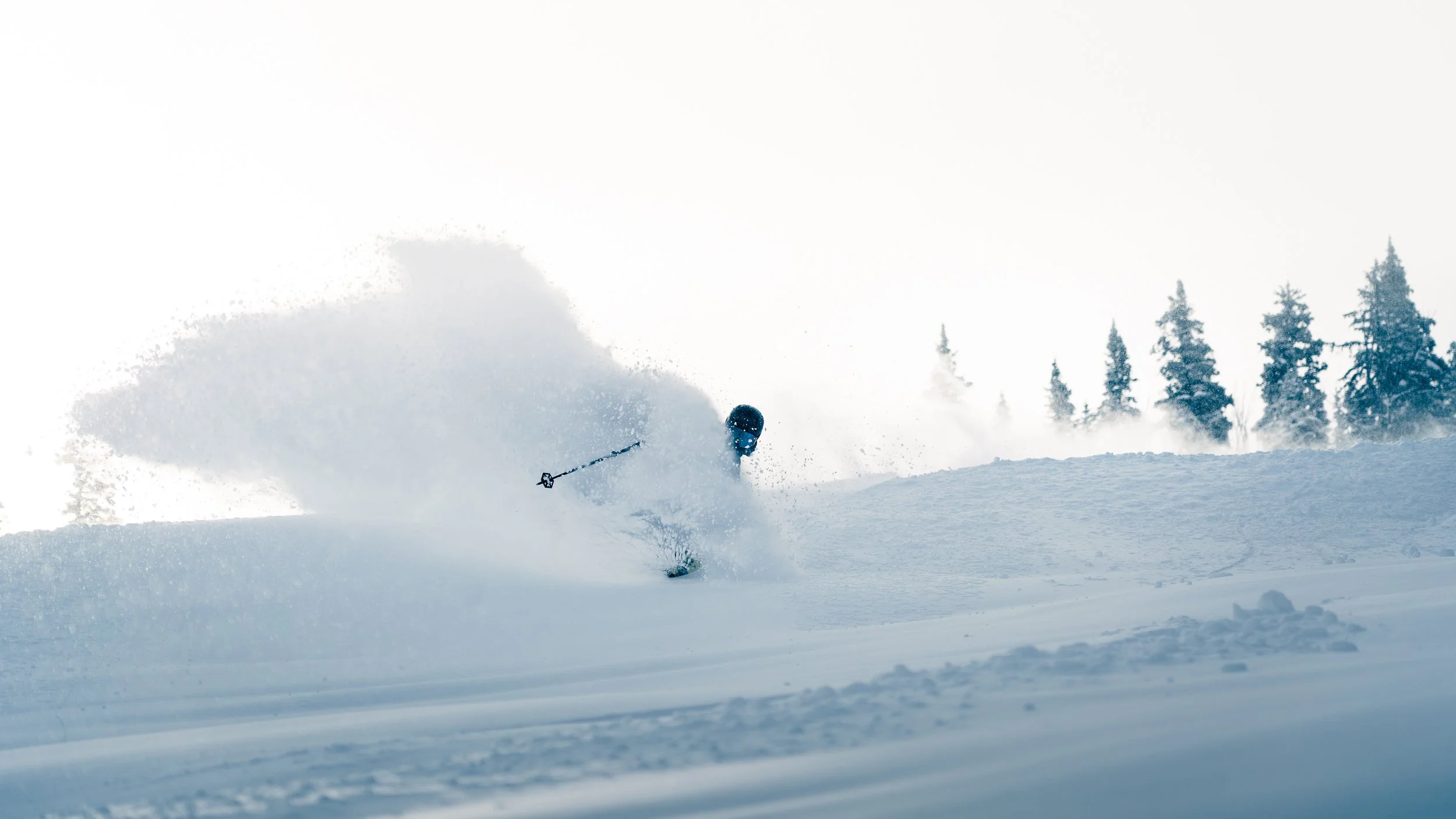 A skier in black gear skiing down a snowy slope, kicking up a spray of snow, with a backdrop of snow-covered trees and a cloudy sky.