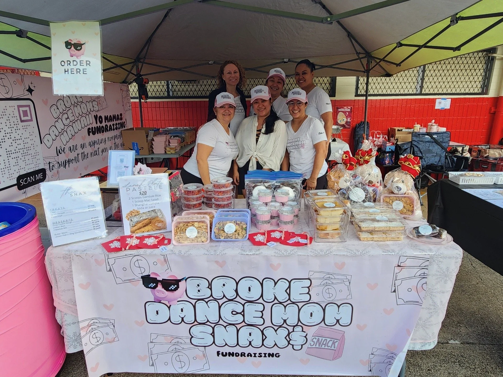 A group of six women standing behind a table at an outdoor fundraising event, with snacks and baked goods for sale, under a tent with colorful panels.