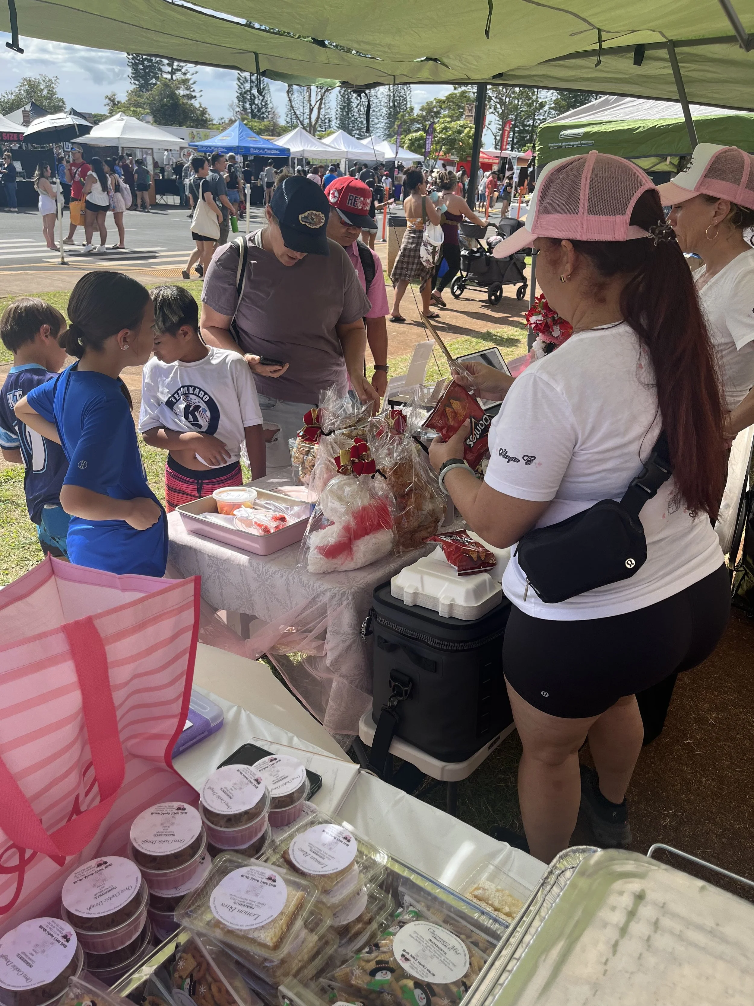 People shopping at an outdoor market stall with desserts, baked goods, and packaged snacks. Several individuals are browsing and engaging with the vendors under a green canopy. Market tents and a crowd of people are visible in the background.