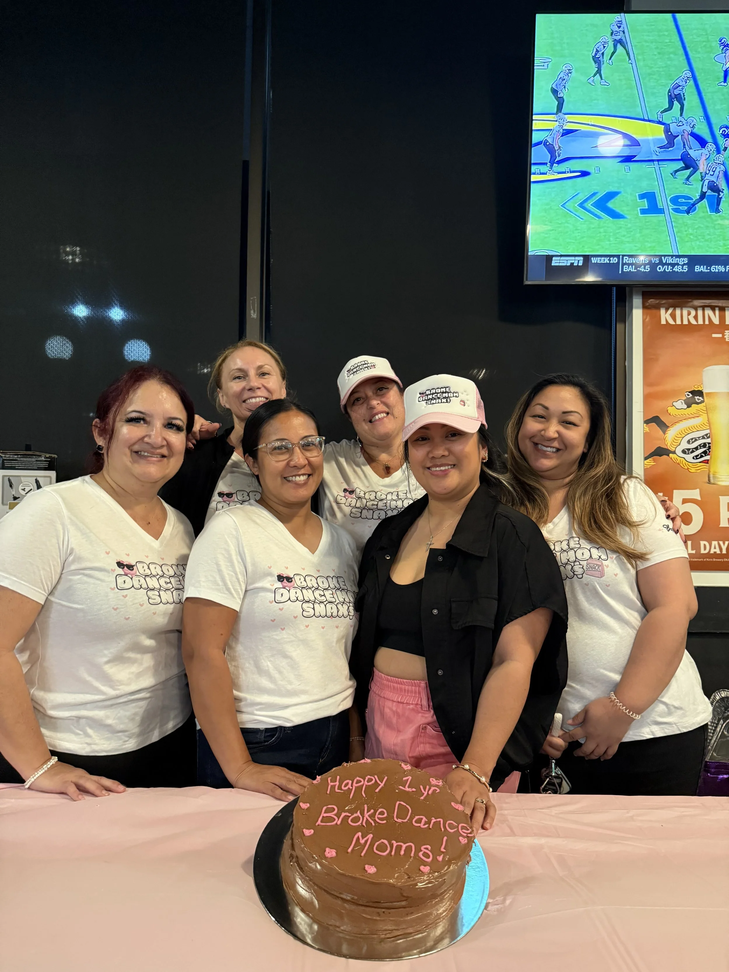 Group of women celebrating a 1-year anniversary with a chocolate cake that has pink icing writing and hearts, in front of a television screen showing a football game.