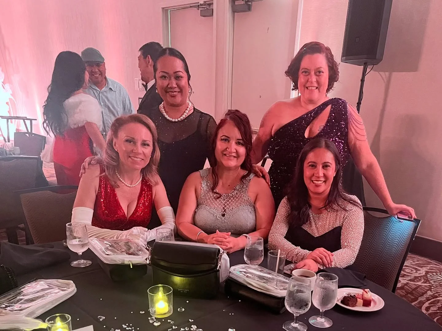 Group of six women dressed in evening attire at a dinner event, sitting and standing around a table with glasses of water, plates with desserts, and a candle.