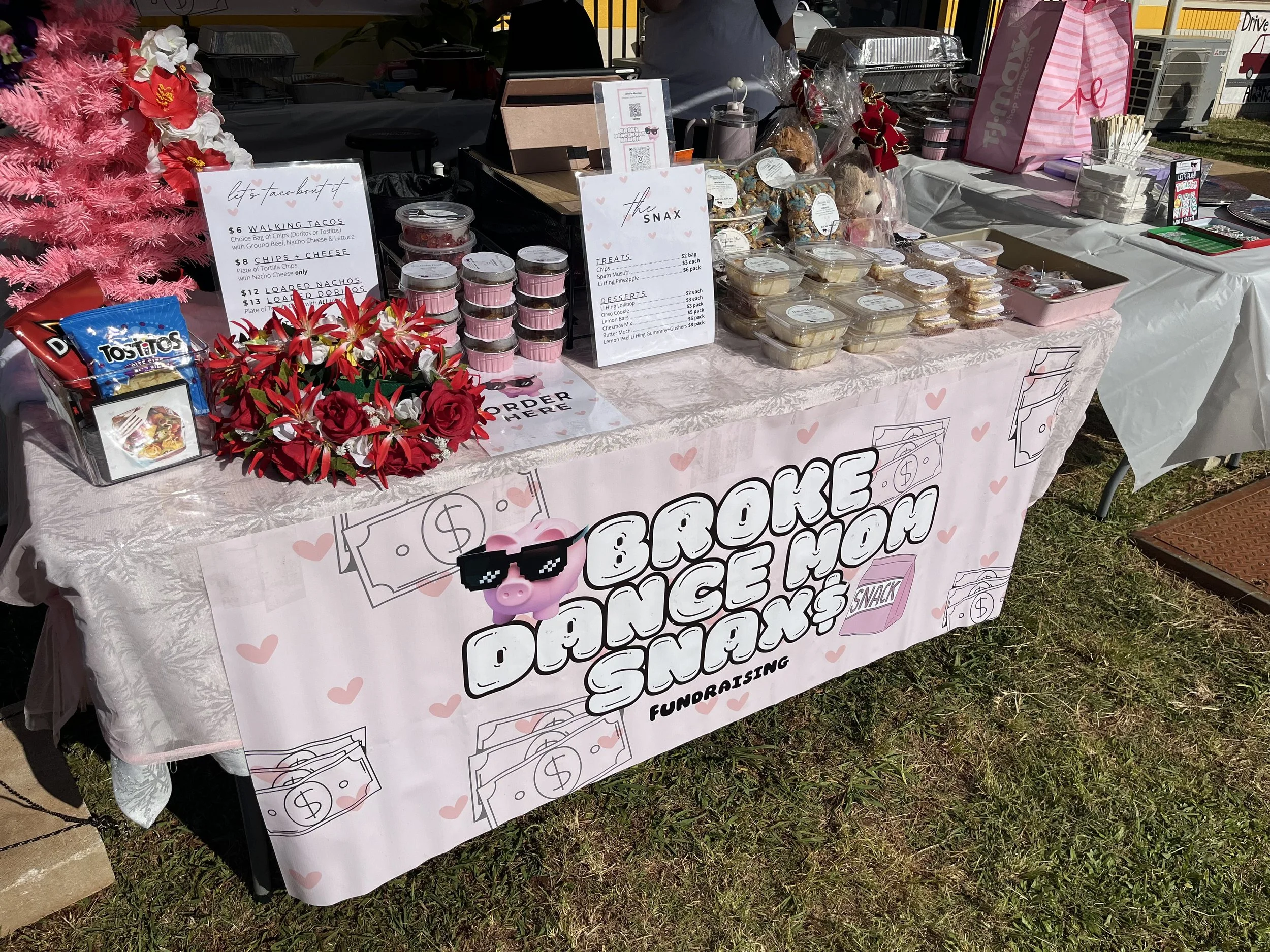 A table at a fundraiser event decorated with a pink tablecloth and pink hearts, selling snacks and baked goods. Signs display food prices and options. Items include cupcakes, cookies, and candy. There is a pink 'Love' shopping bag, artificial pink fl