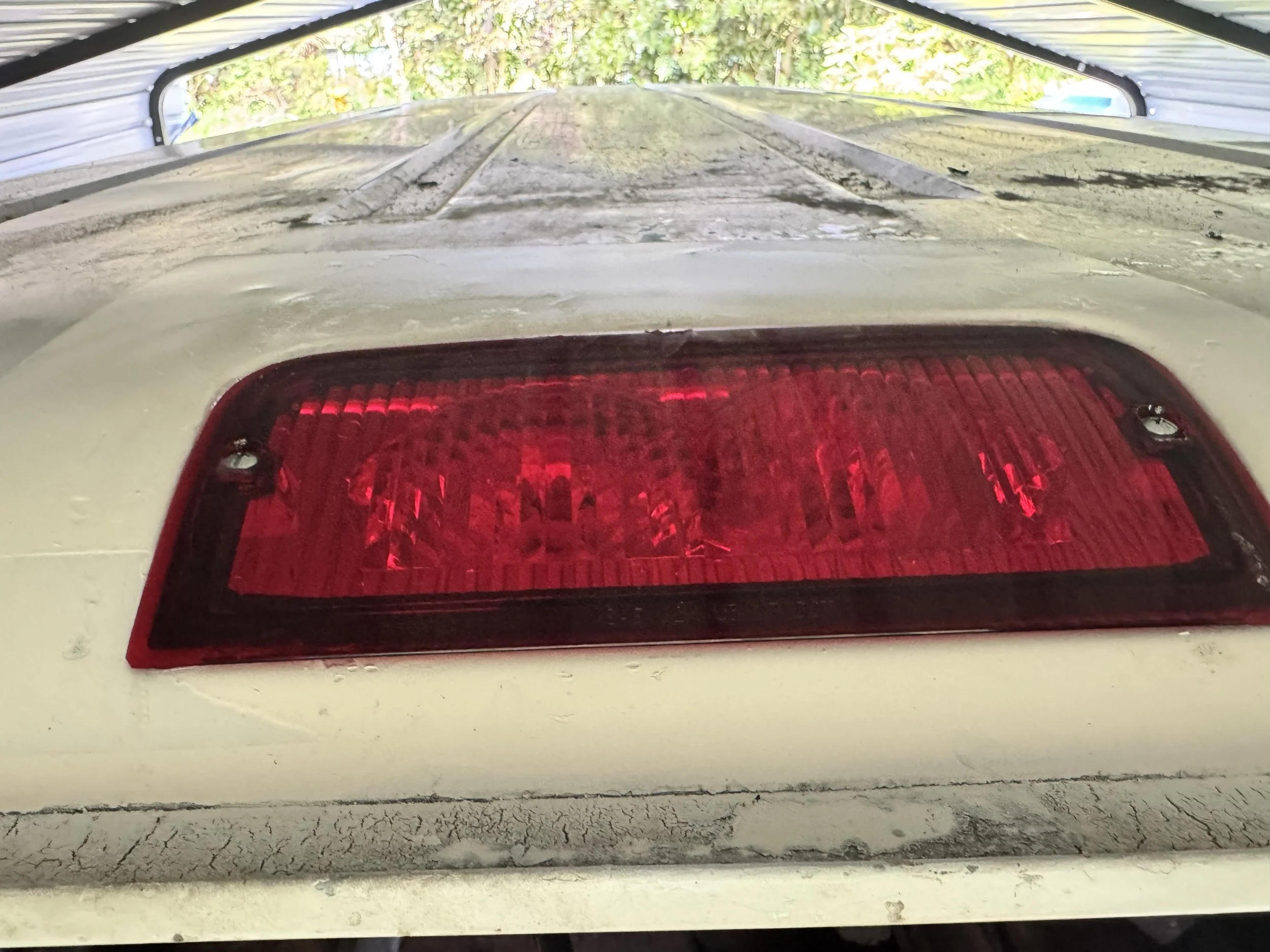 Close-up of a car's taillight mounted on an off-white bumper, with a weathered surface and a crack at the bottom, viewed from behind during daytime.