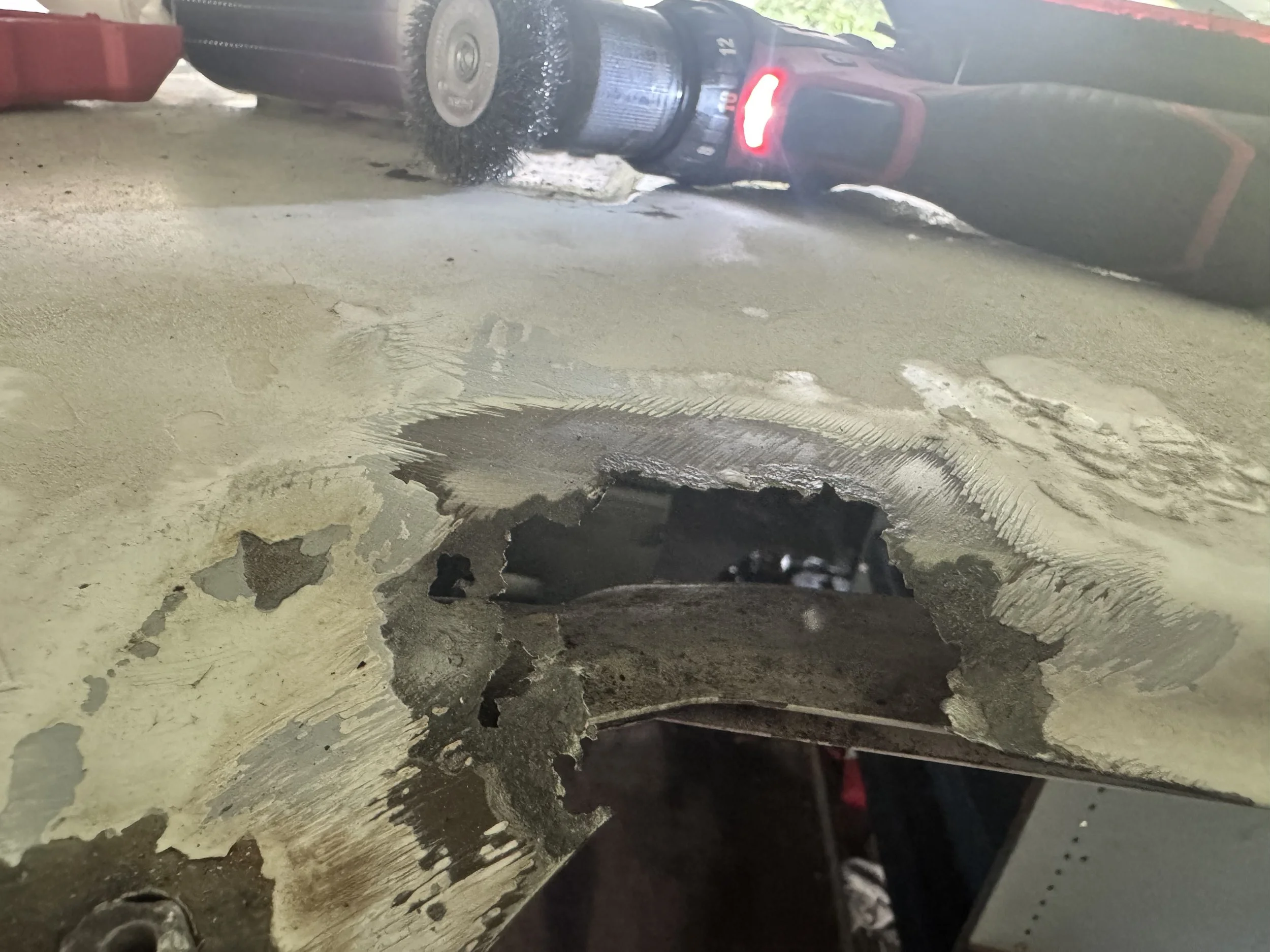 Close-up of a damaged car door with peeling paint and rust, showing a hole in the metal and scratches, with a reflection of a garage or workshop environment.