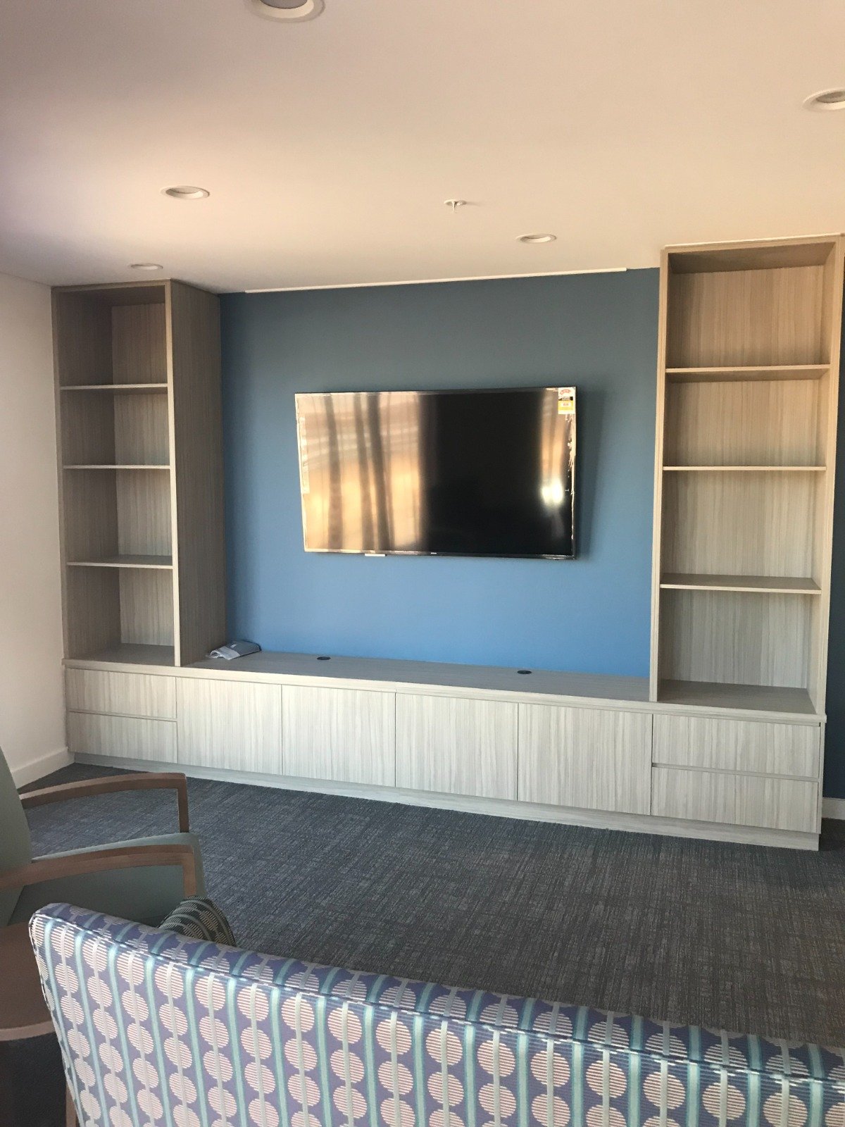 Living room with wall-mounted flat-screen TV, built-in shelves on both sides, a low cabinet below, a patterned armchair, and a blue accent wall.
