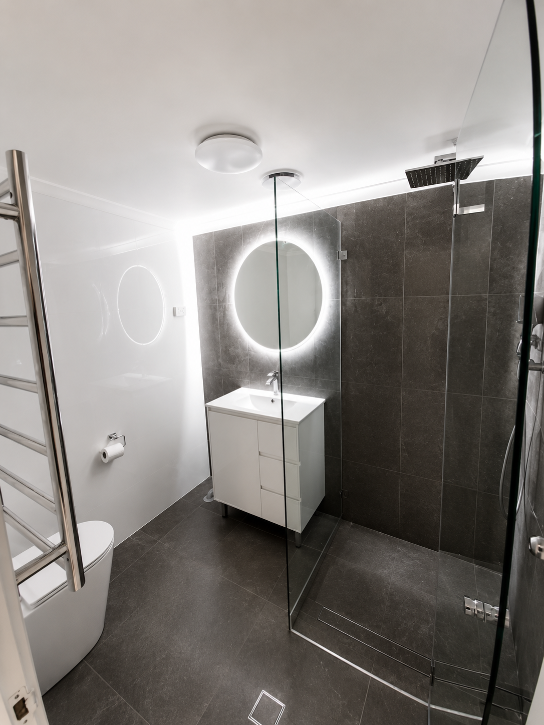 Modern bathroom with dark tiled walls and floor, white vanity with oval mirror backlit by LED lighting, glass shower enclosure, and sleek fixtures.