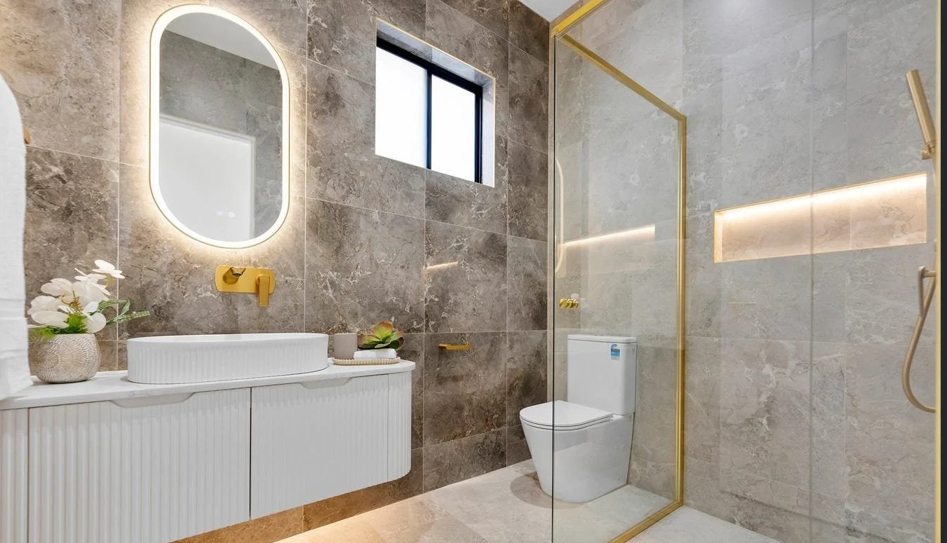 Modern bathroom with gray marble tiles, a white Vanity with a basin sink, golden fixtures, a decorative plant, a mirror with backlighting, a small window, and a separate enclosed shower area with glass and gold trim.