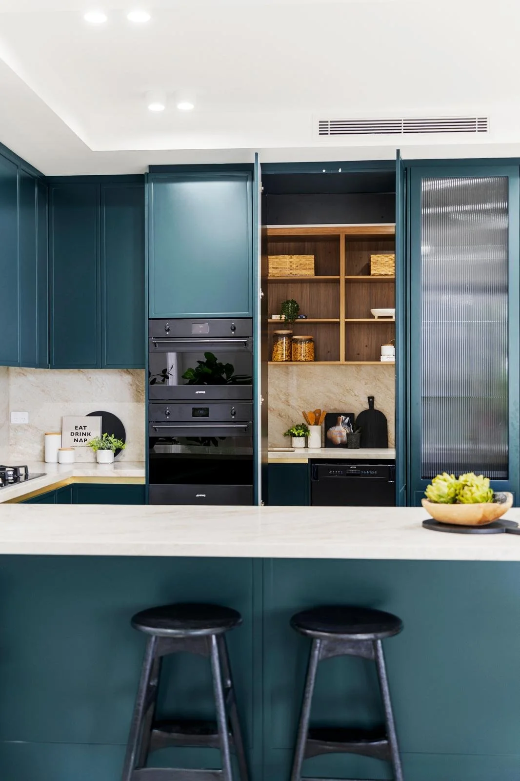 Modern kitchen with blue cabinets, marble countertops, open shelving with baskets and jars, and two black stools at a marble island.