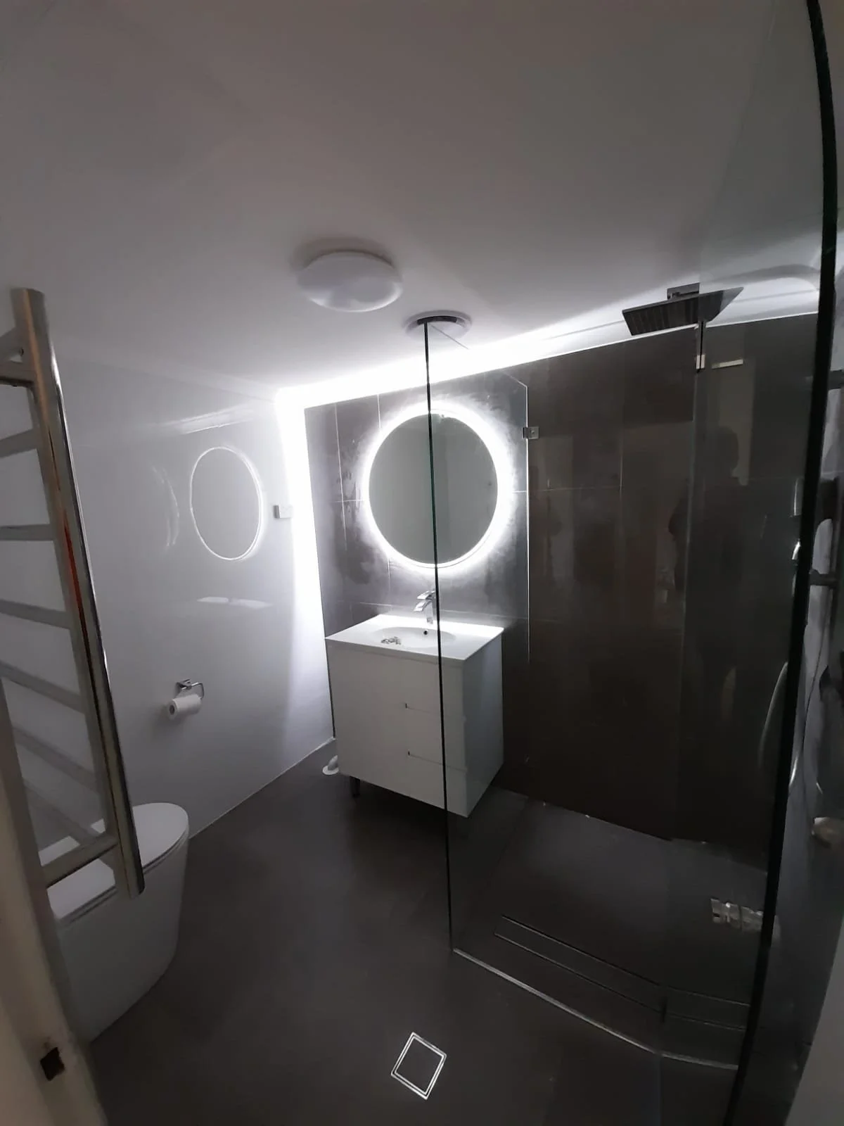 Modern bathroom with a white sink and vanity, echoing white walls, a round backlit mirror, toilet, and a glass shower enclosure.