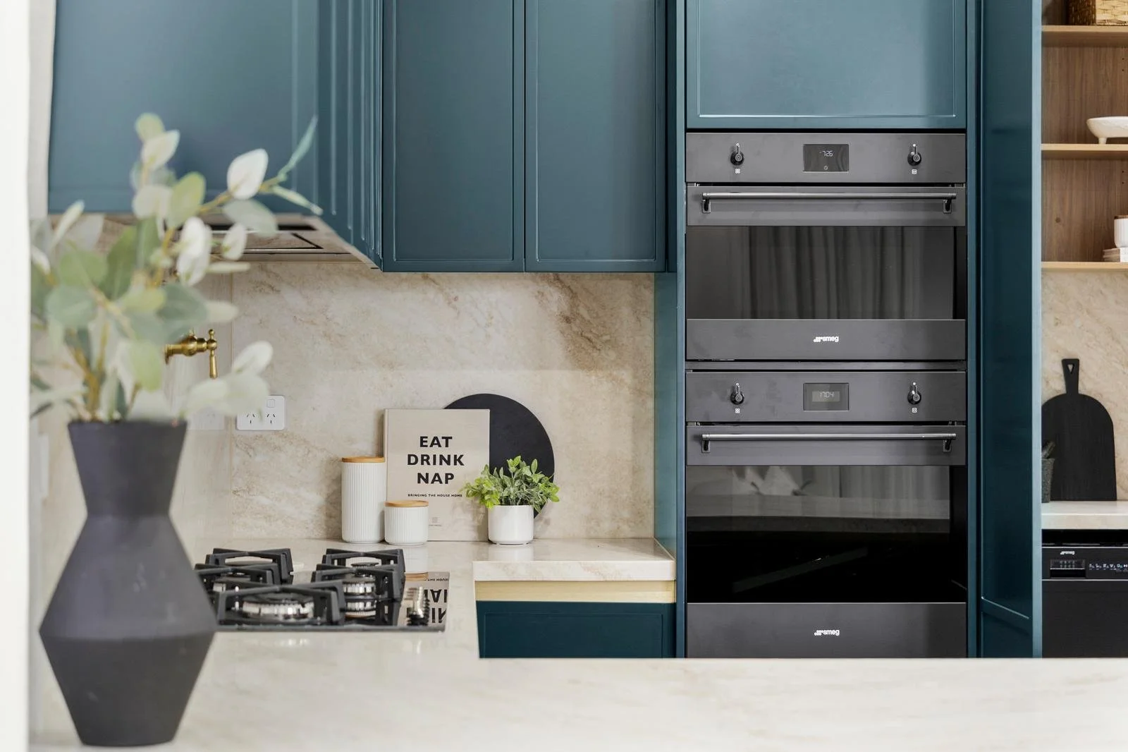 Modern kitchen with blue cabinets, black oven, beige marble backsplash, and decorative items on the counter including a plant, a sign that reads 'Eat Drink Nap,' and containers.