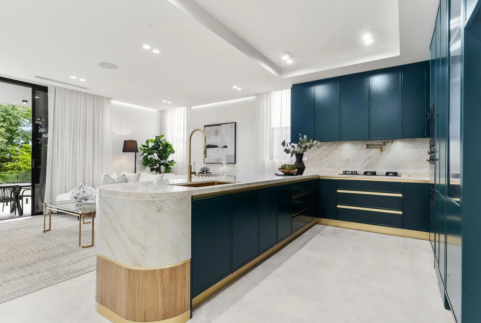 Modern kitchen with dark teal cabinets, cream marble countertops, and a marble backsplash. Open living space with white curtains, plants, and a dining area with a glass sliding door leading outside.
