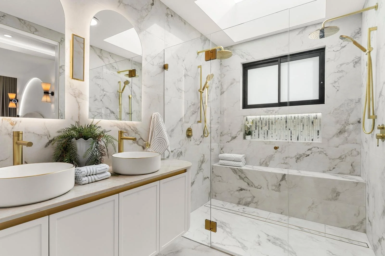 Modern bathroom with marble walls and floors, dual vessel sinks on a vanity with a plant, gold fixtures, a glass shower with gold hardware, and a window with frosted glass.