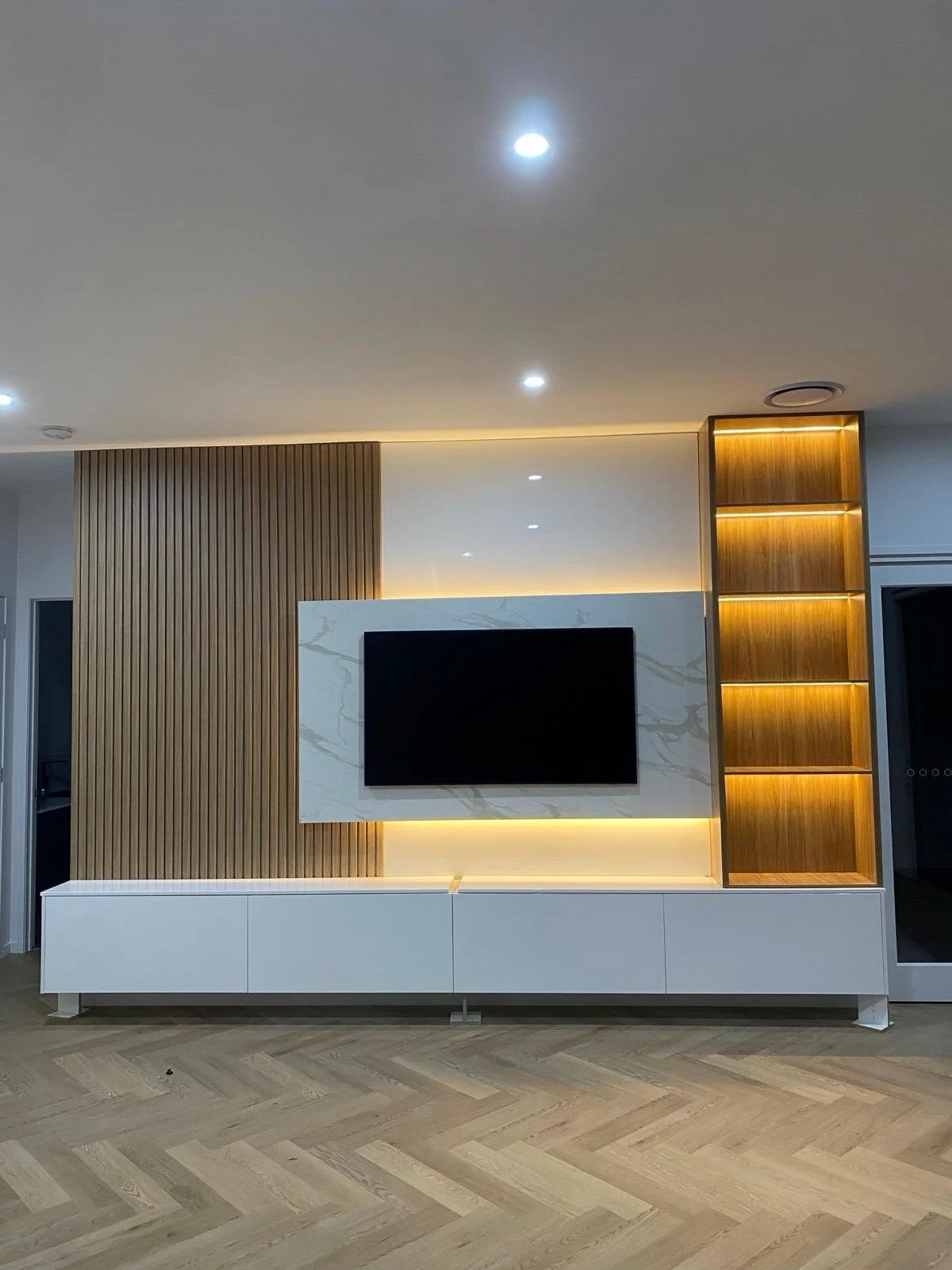 Modern living room wall with a flat-screen TV mounted on a white marble panel, flanked by wooden panels with vertical slats and backlit shelves, and a white media console below.