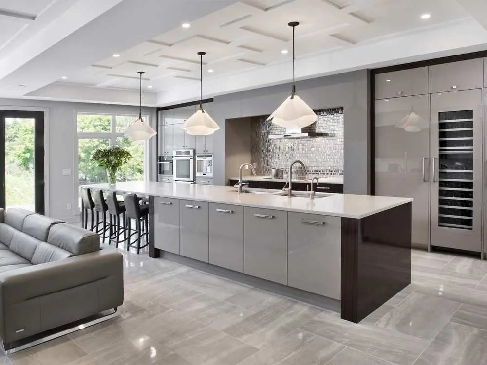 Modern kitchen with white island, black chairs, stainless steel appliances, gray cabinets, large window with greenery, pendant lights, and tiled flooring.