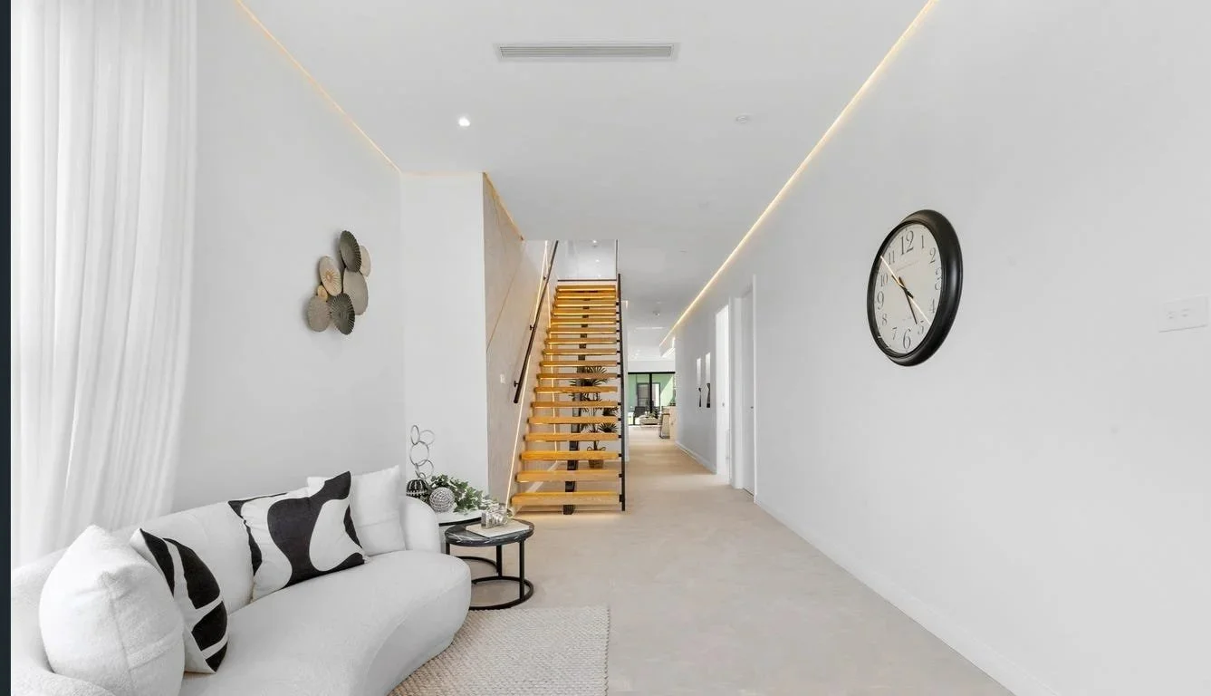 Modern, minimalist home interior with white walls, beige flooring, a white sofa with black and white pillows, a small black side table with decorative items, a wooden staircase with black railing, and a large black wall clock.