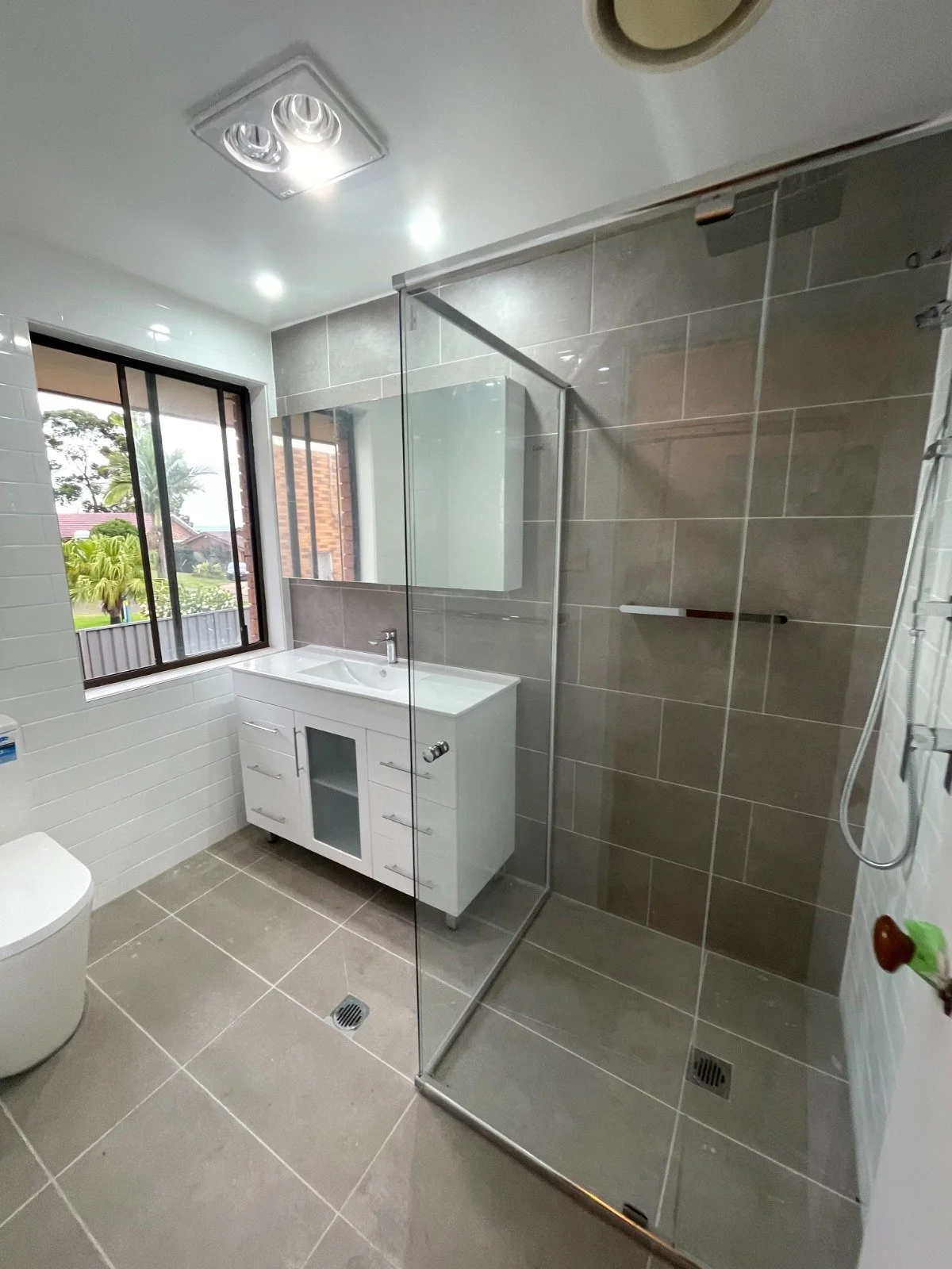 Modern bathroom with a large window, white vanity with sink, and a glass-enclosed shower with brown tiles.